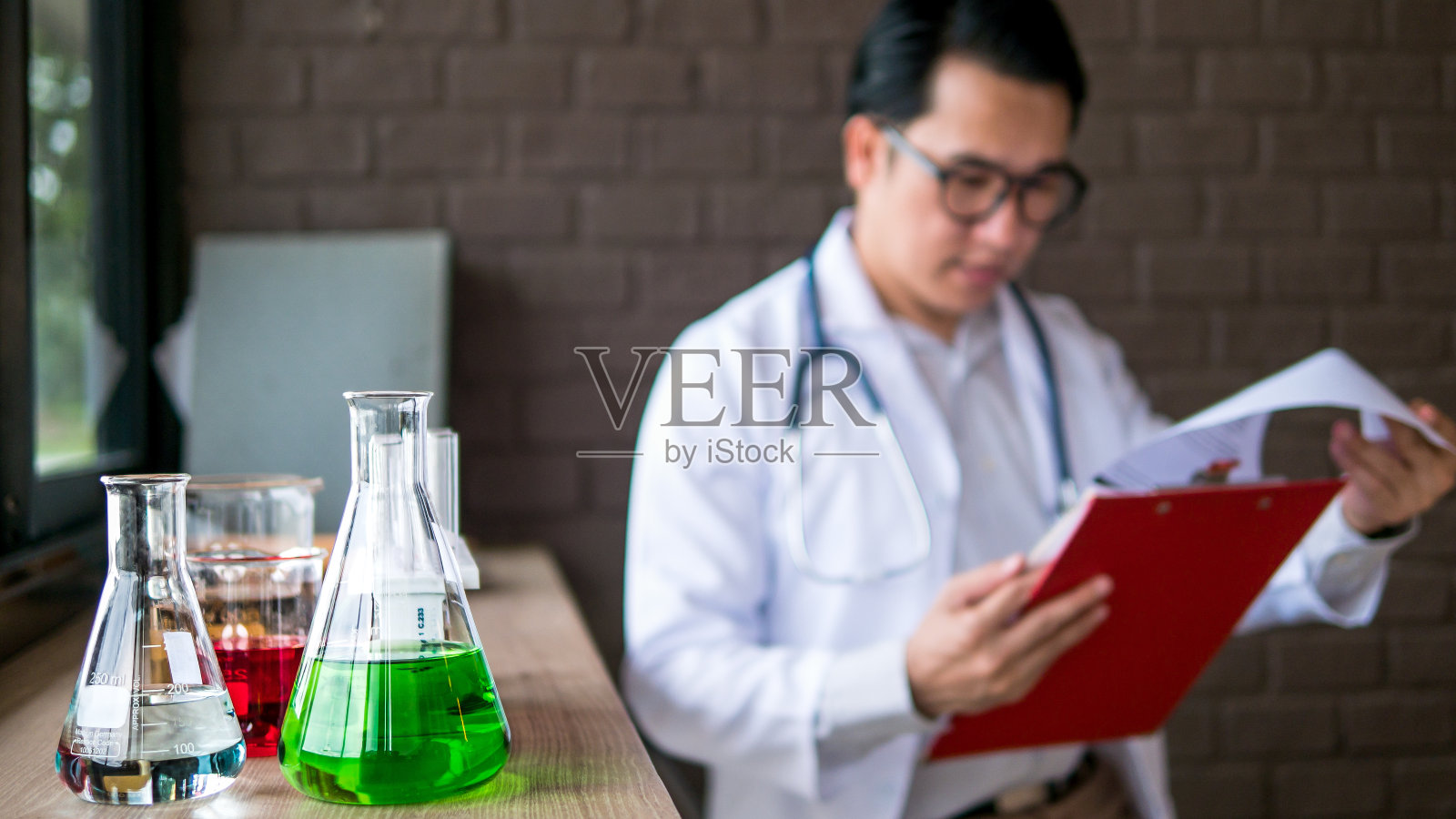 医疗工作的代理机构。选择性聚焦图像的实验室玻璃器皿与彩色液体化学和模糊的背景年轻的亚洲医生阅读报告文件。照片摄影图片