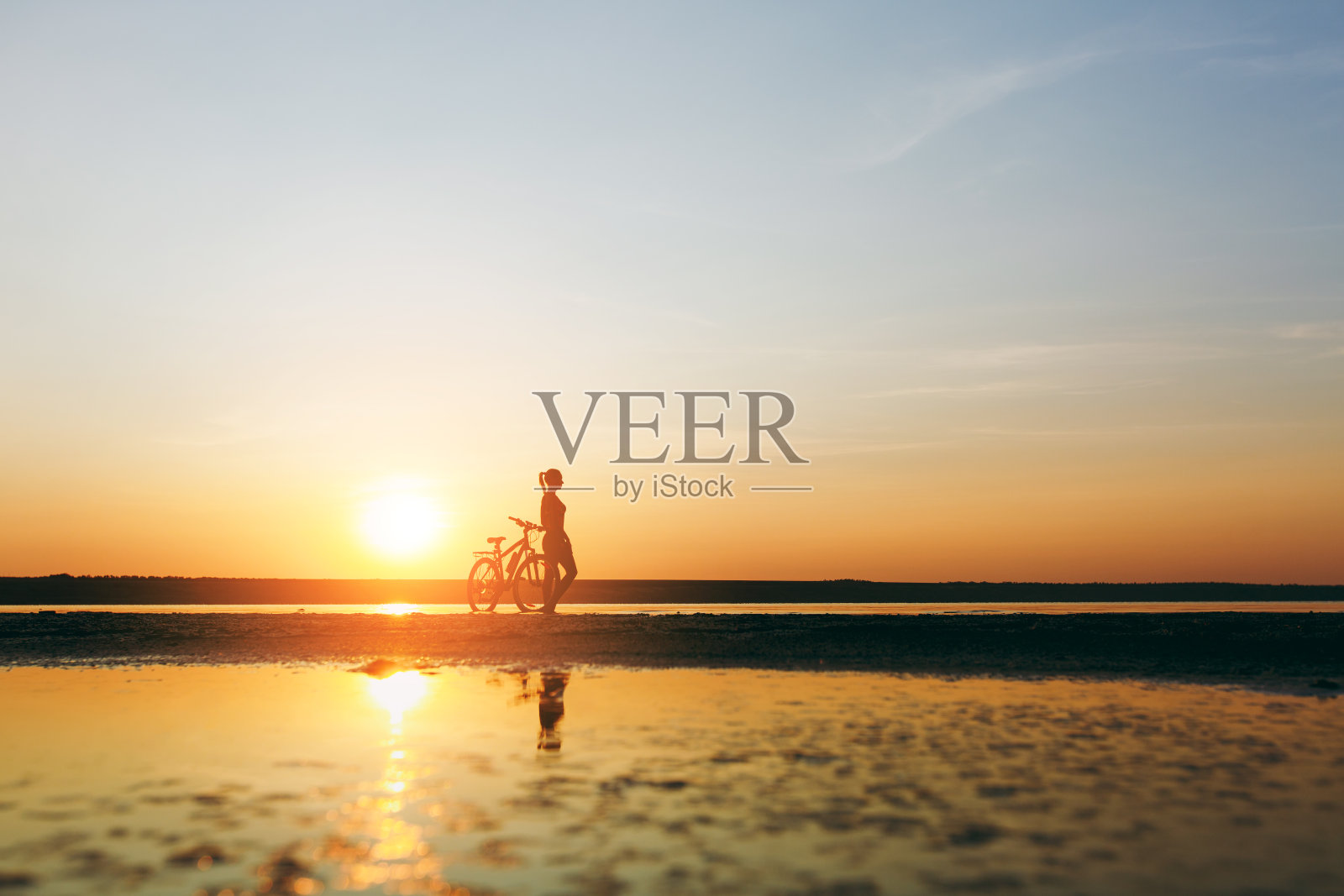 一个运动女孩的剪影在一个温暖的夏天日落在水附近的自行车。健康的概念。天空背景照片摄影图片