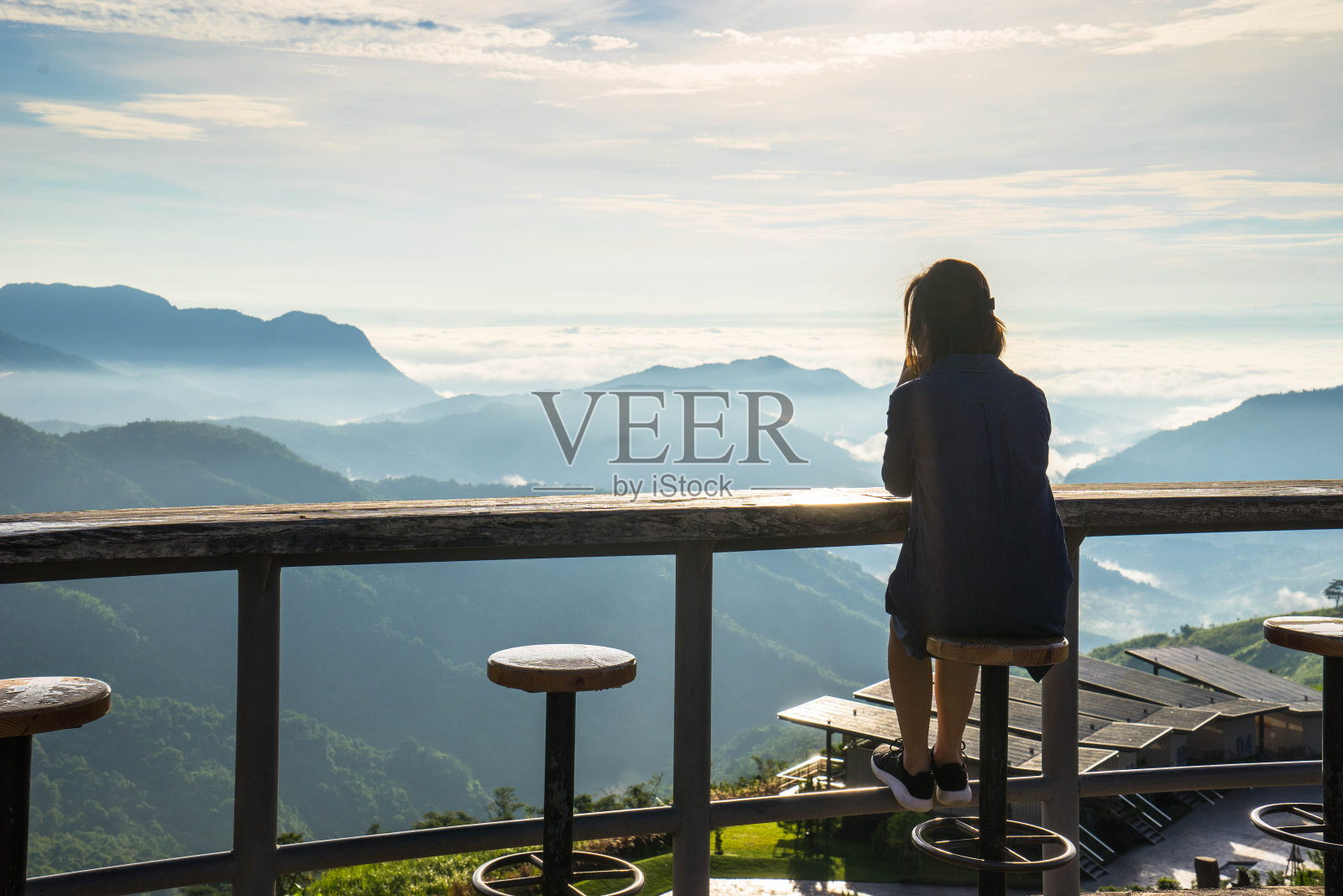 女人坐在凳子上，看着日出下的山脉和薄雾的全景照片摄影图片