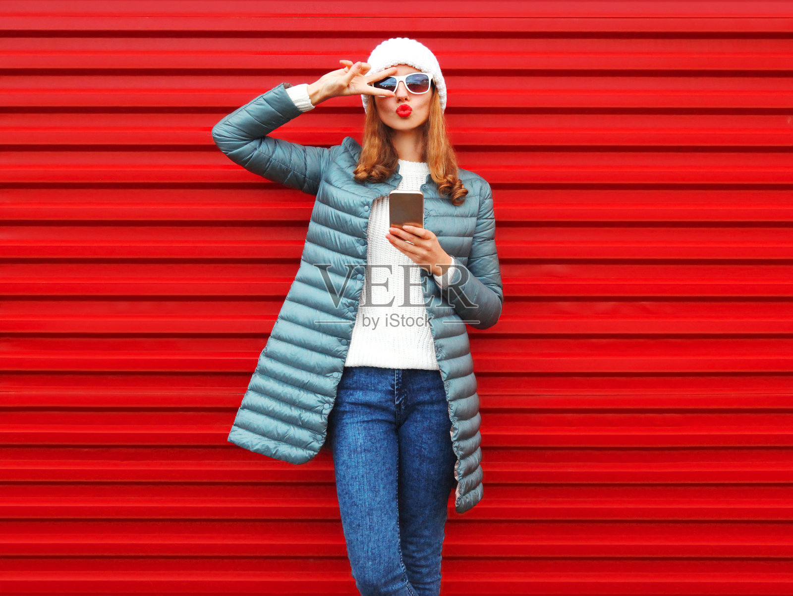 时尚美女吹着嘴唇，在城市的红色背景下使用智能手机照片摄影图片