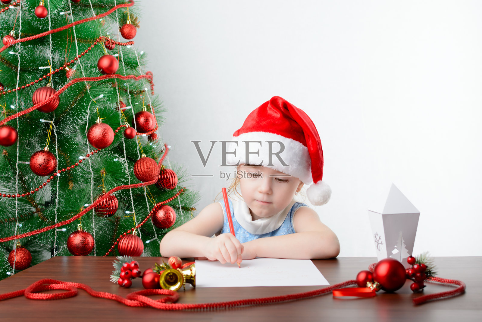 给圣诞老人写信的概念。可爱的小女孩拿着一支笔坐在圣诞树旁的桌子旁照片摄影图片