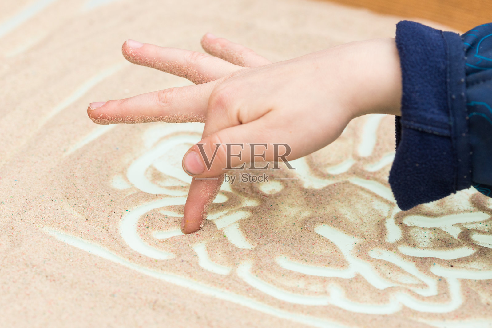 绘画、创意、手工概念。穿着牛仔衬衫的小孩的小臂的近处，他用手指在被沙子覆盖的白色表面上画着线条和卷曲照片摄影图片