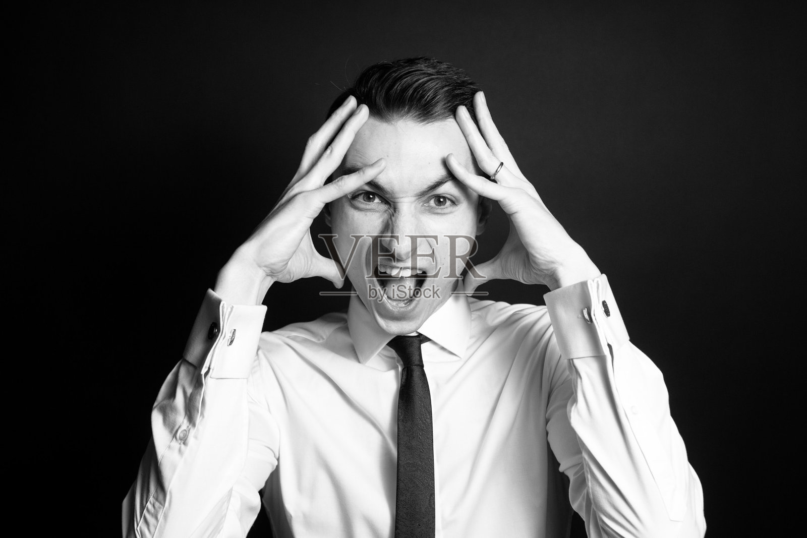 黑白照片上，一个男人穿着白衬衫，打着黑领带，双手抱头，对着镜头尖叫。照片摄影图片