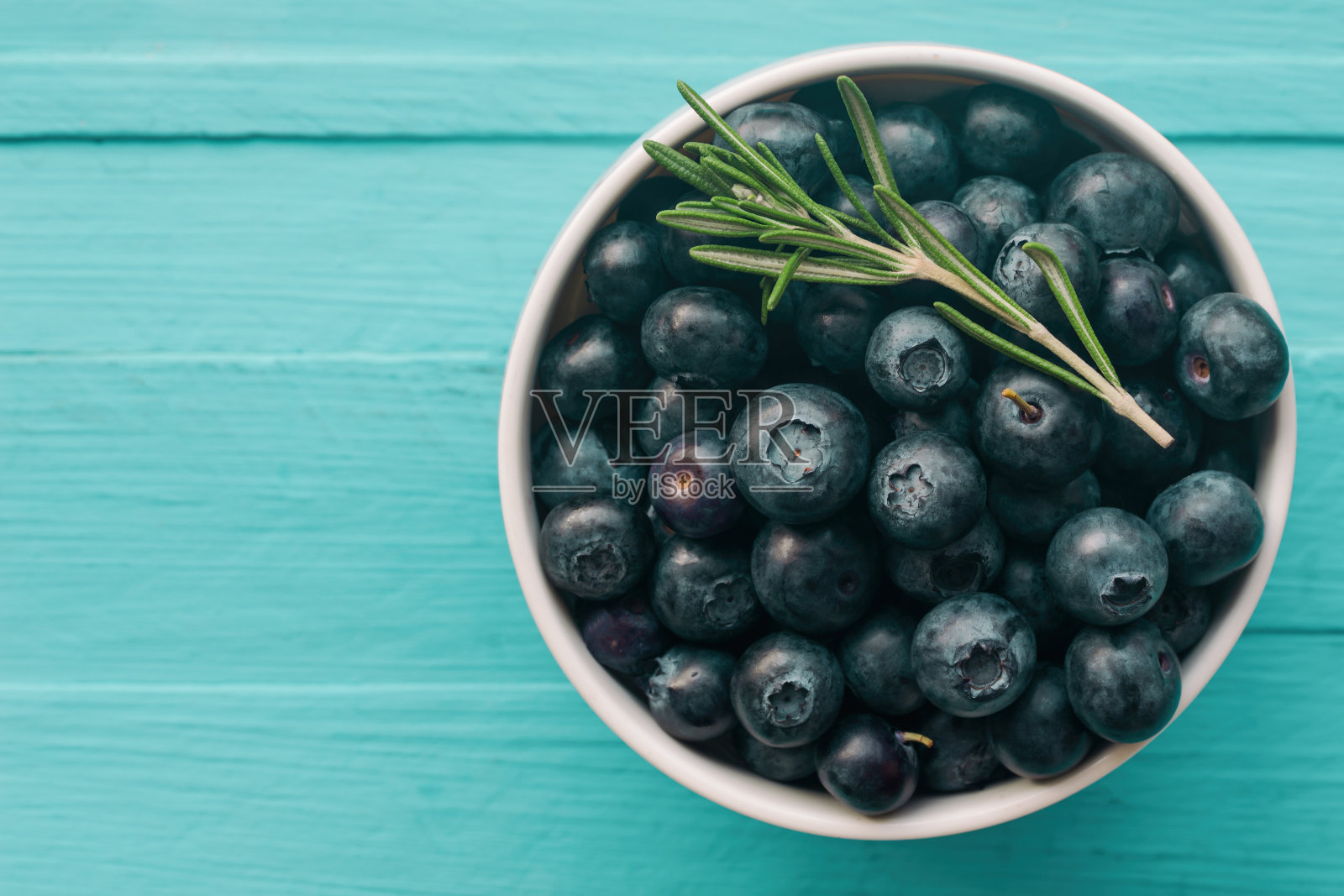 新鲜成熟的野生蓝莓在白色的碗里，在俯视图的蓝色木桌上平躺着，背景是复制空间。蓝莓是抗氧化食品和维生素c，是健康美味的水果概念。照片摄影图片