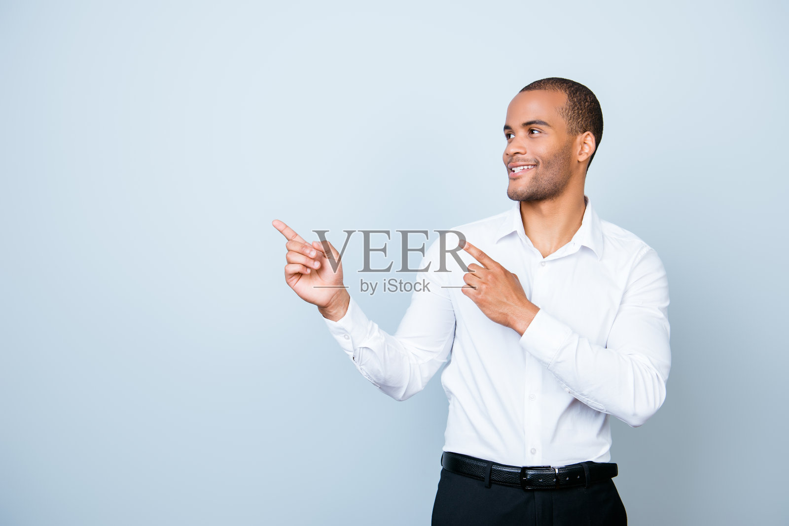 在纯浅蓝色的背景上，一个年轻的、快乐的、成功的非洲企业家面带微笑，穿着正装，用手指指着一个版面照片摄影图片