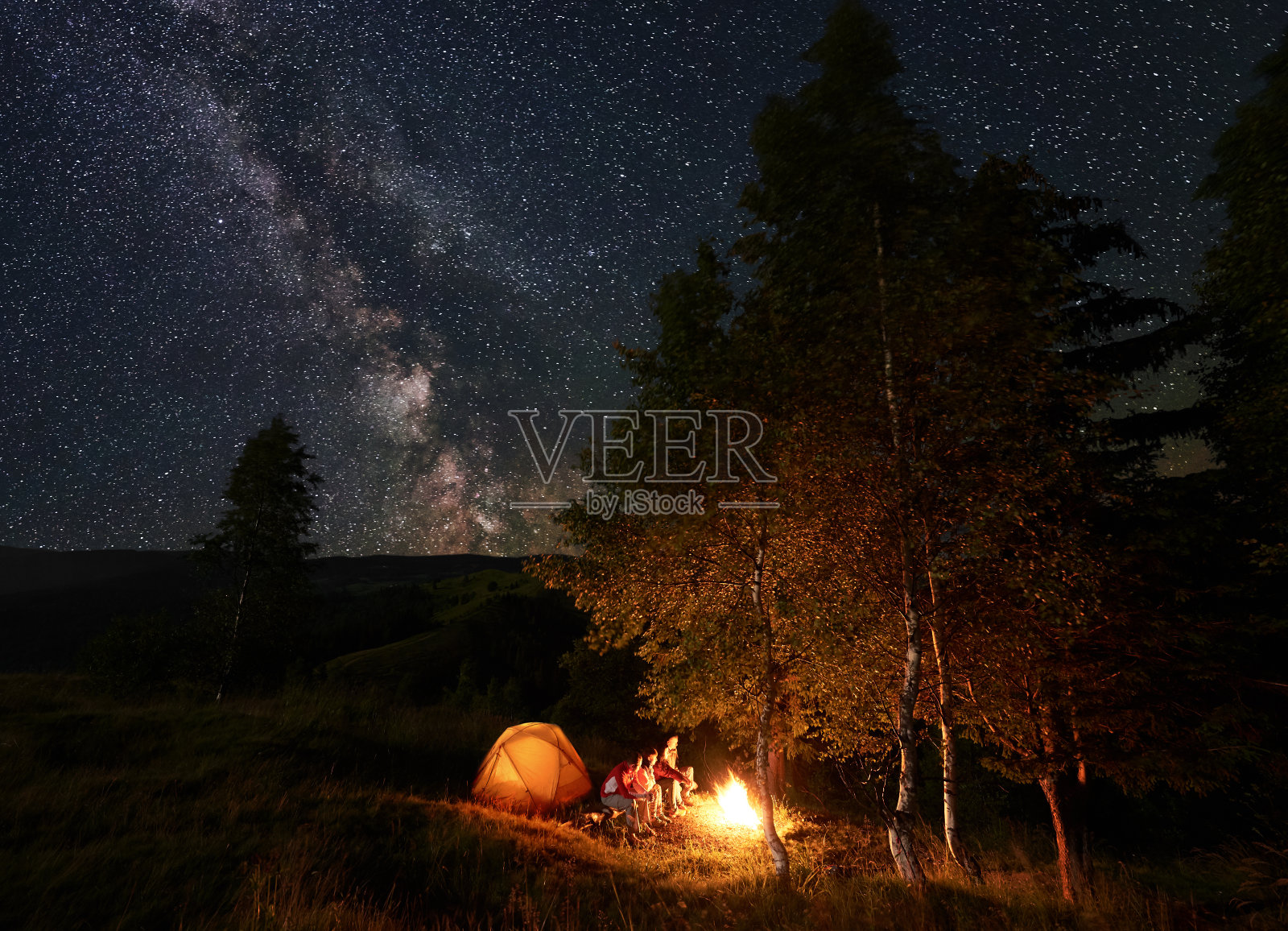 在星空下，一个年轻人坐在篝火旁的圆木上，在树木中露营，靠近帐篷照片摄影图片