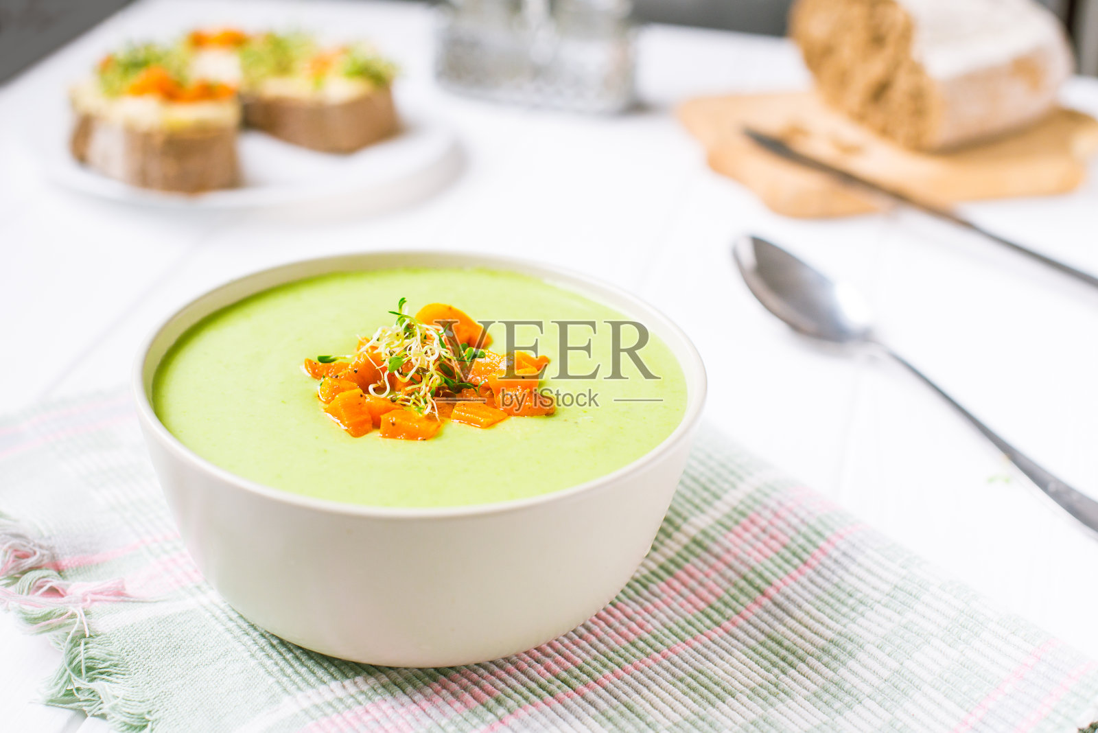 白色的木桌上放着一碗绿豌豆奶油汤，还有烤胡萝卜和微绿芽。健康食品，素食饮食理念。选择性聚焦，文本空间。照片摄影图片