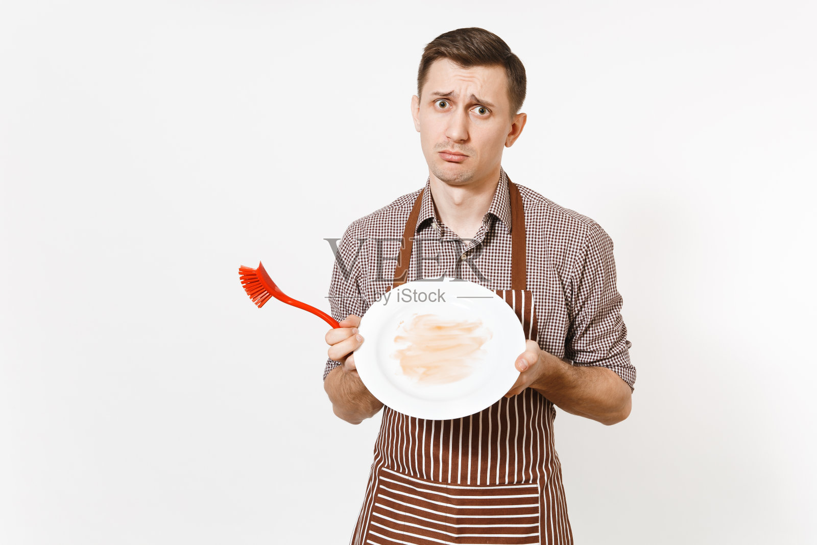 有趣的男厨师在条纹棕色围裙，衬衫拿洗白色圆形空脏盘子与红刷清洗盘子孤立在白色的背景。男管家、家务工人或家庭佣工照片摄影图片