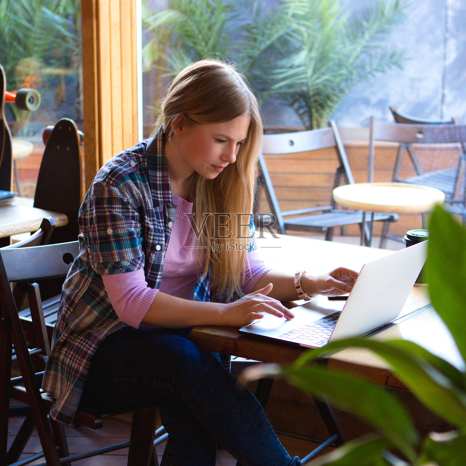金发女孩坐在舒适时髦的咖啡馆里拿着笔记本电脑和咖啡杯。在上网本上打字的女性自由职业者。照片摄影图片