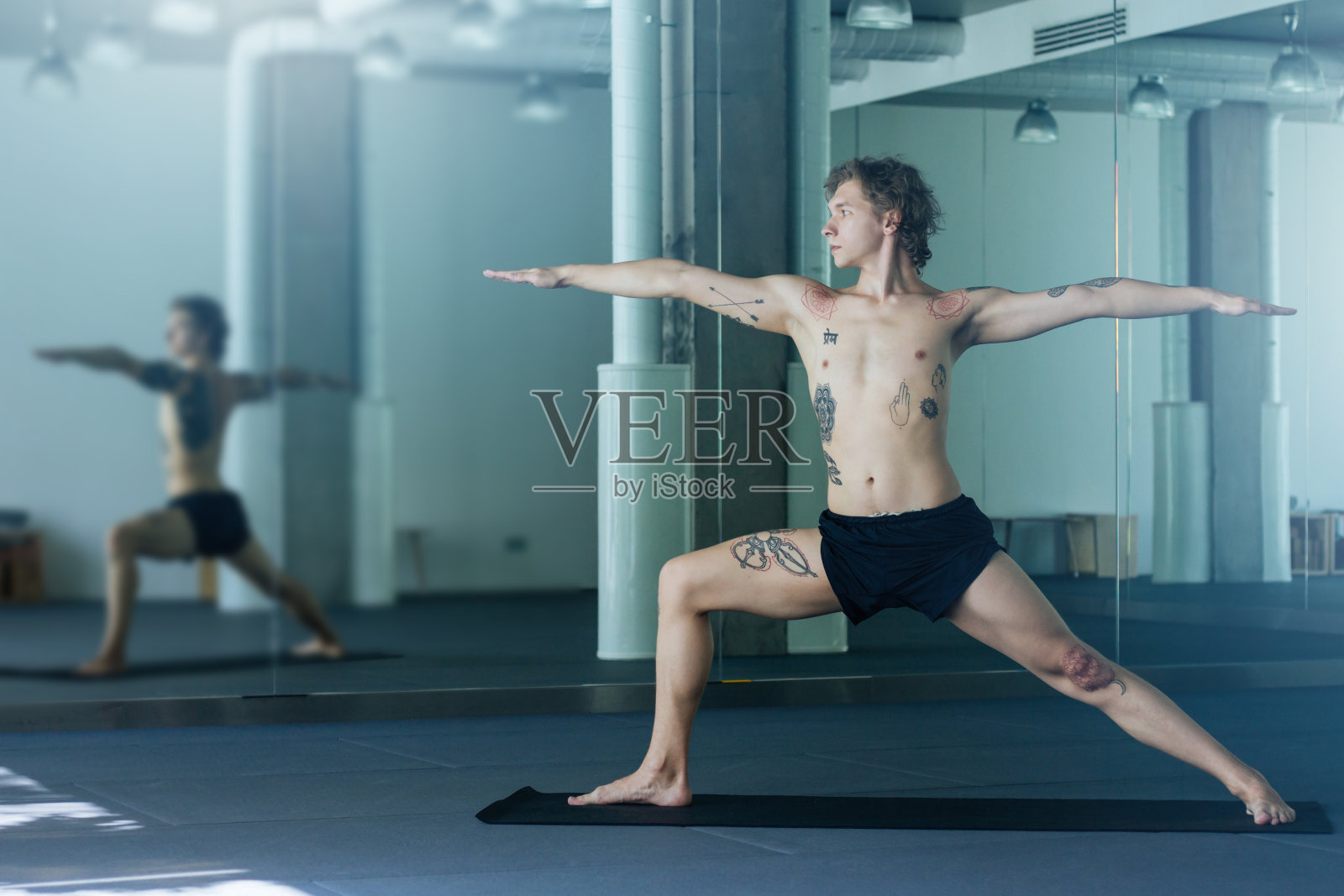 英俊的纹身男子练习瑜伽。年轻的男老师在教室里做武士式，在黑色垫子上做Virabhadrasana。瑜伽大师的概念照片摄影图片