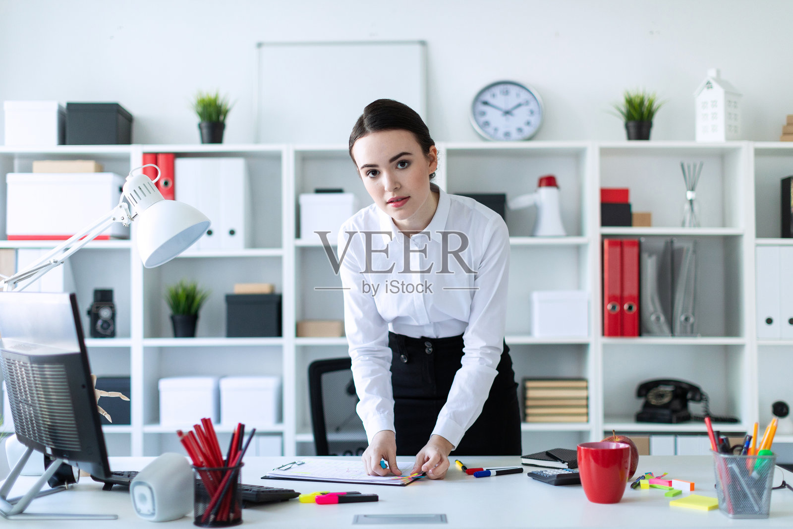 一个年轻的女孩站在办公室靠近桌子的地方，用胶水把文件粘在一起。照片摄影图片