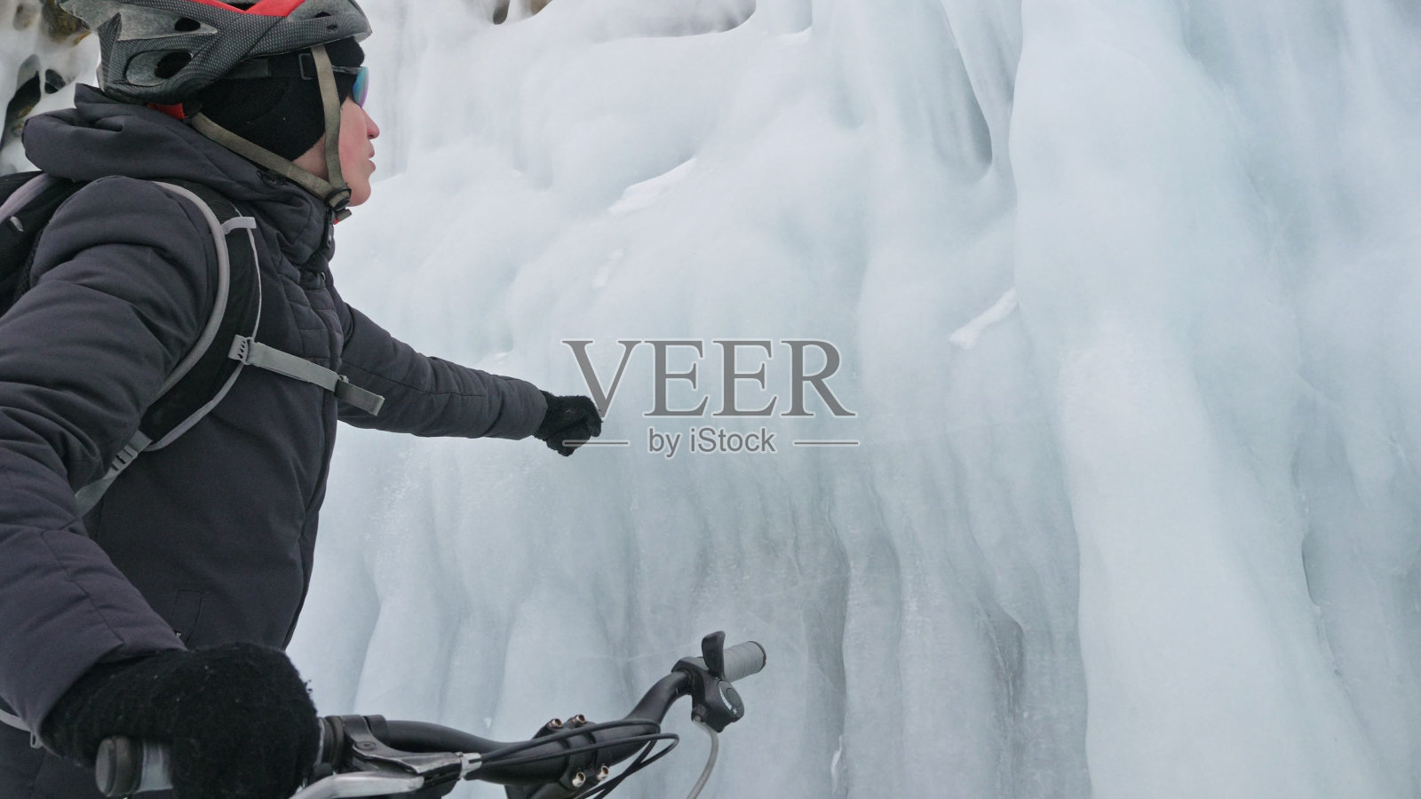 在冰洞附近，一个人在自行车旁边行走。有冰洞和冰柱的岩石非常漂亮。骑自行车的人穿着灰色羽绒服，背着背包，戴着头盔。旅行者正在骑自行车。照片摄影图片