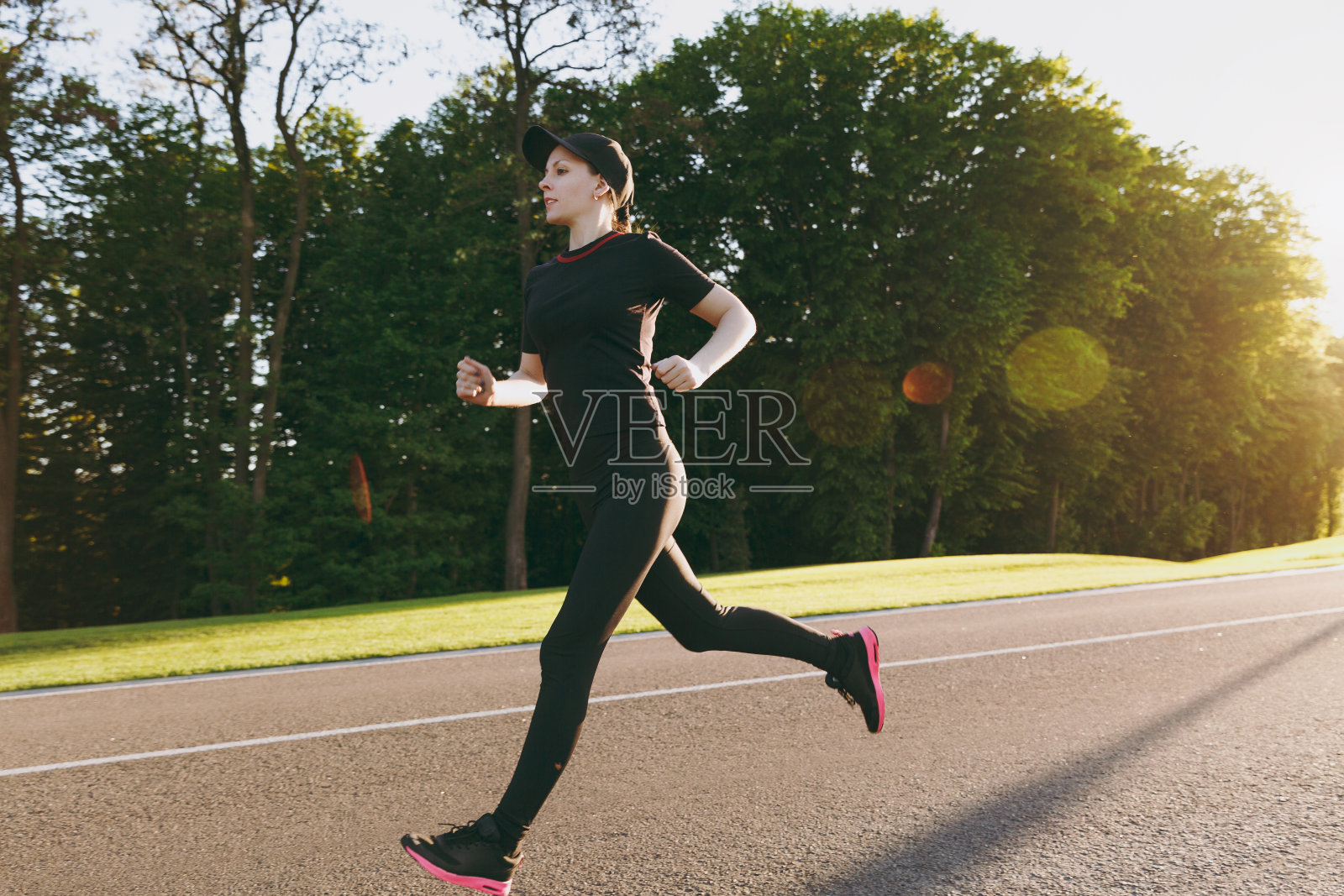 年轻的运动女孩穿着黑色制服和帽子训练，做运动，跑步，在春天或夏天阳光明媚的日子在城市公园户外的道路上直视。健身、健康的生活理念。照片摄影图片