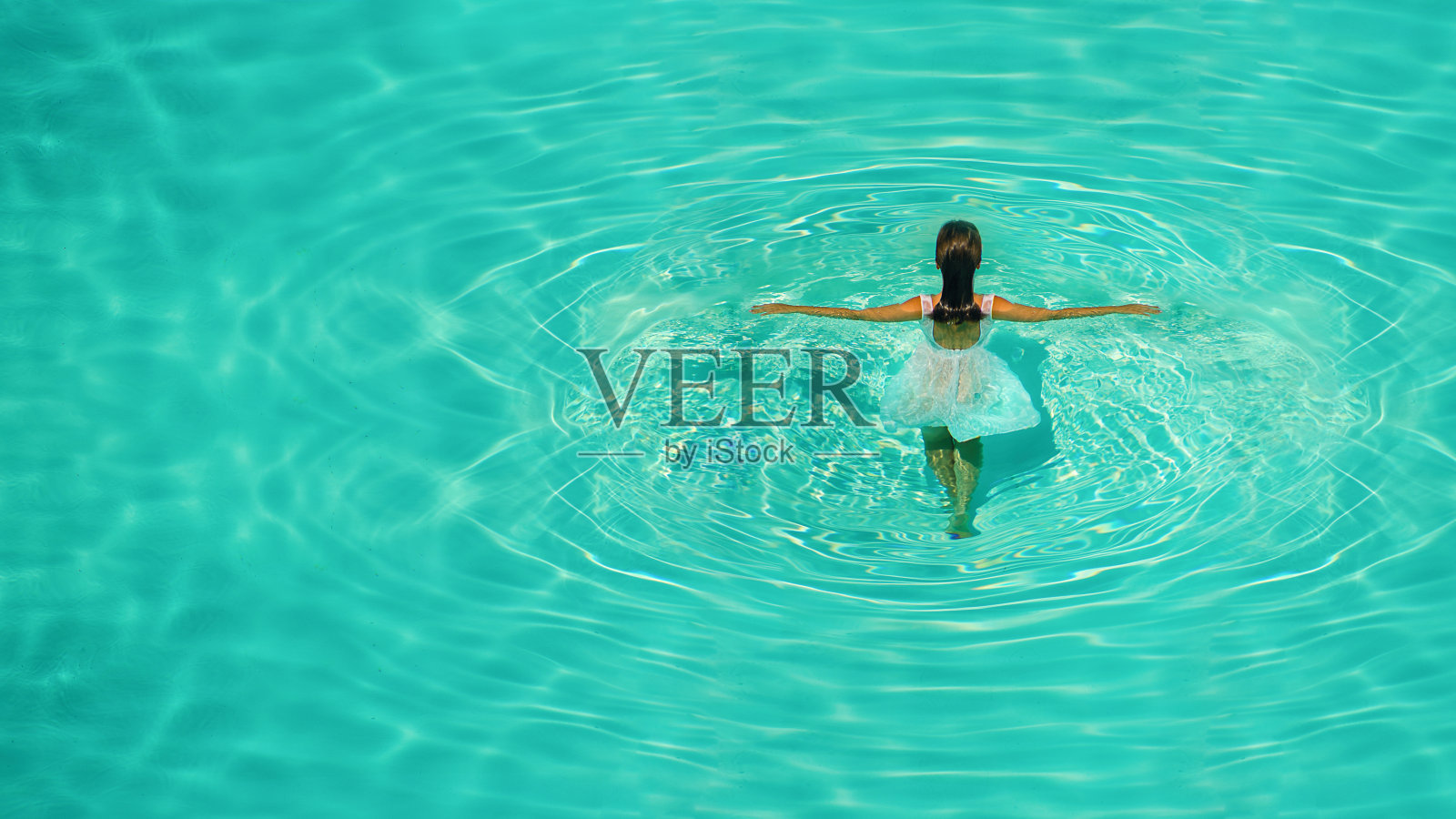 在一个晴朗阳光明媚的下午，穿着白色连衣裙的女孩在蓝色的水池里。照片摄影图片