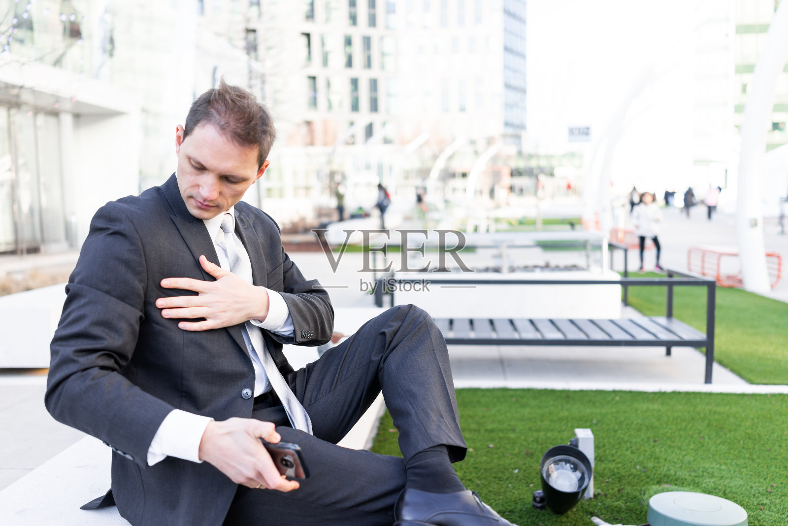 在城市绿地公园的长椅上，一位年轻的商人正穿着西装，带着领带往下看照片摄影图片