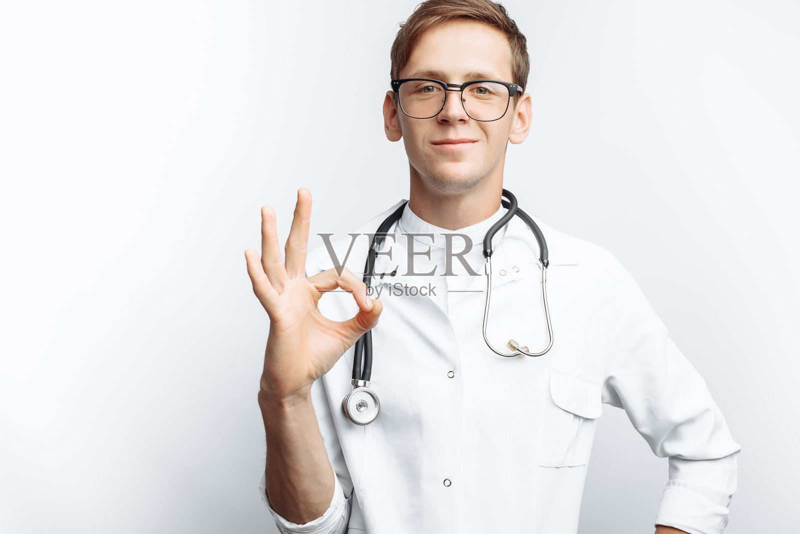 一个年轻医生的肖像，在白色的背景上，显示了手势，好吧，实习生在工作室，脖子上有一个听诊器，用于广告，文字插入照片摄影图片