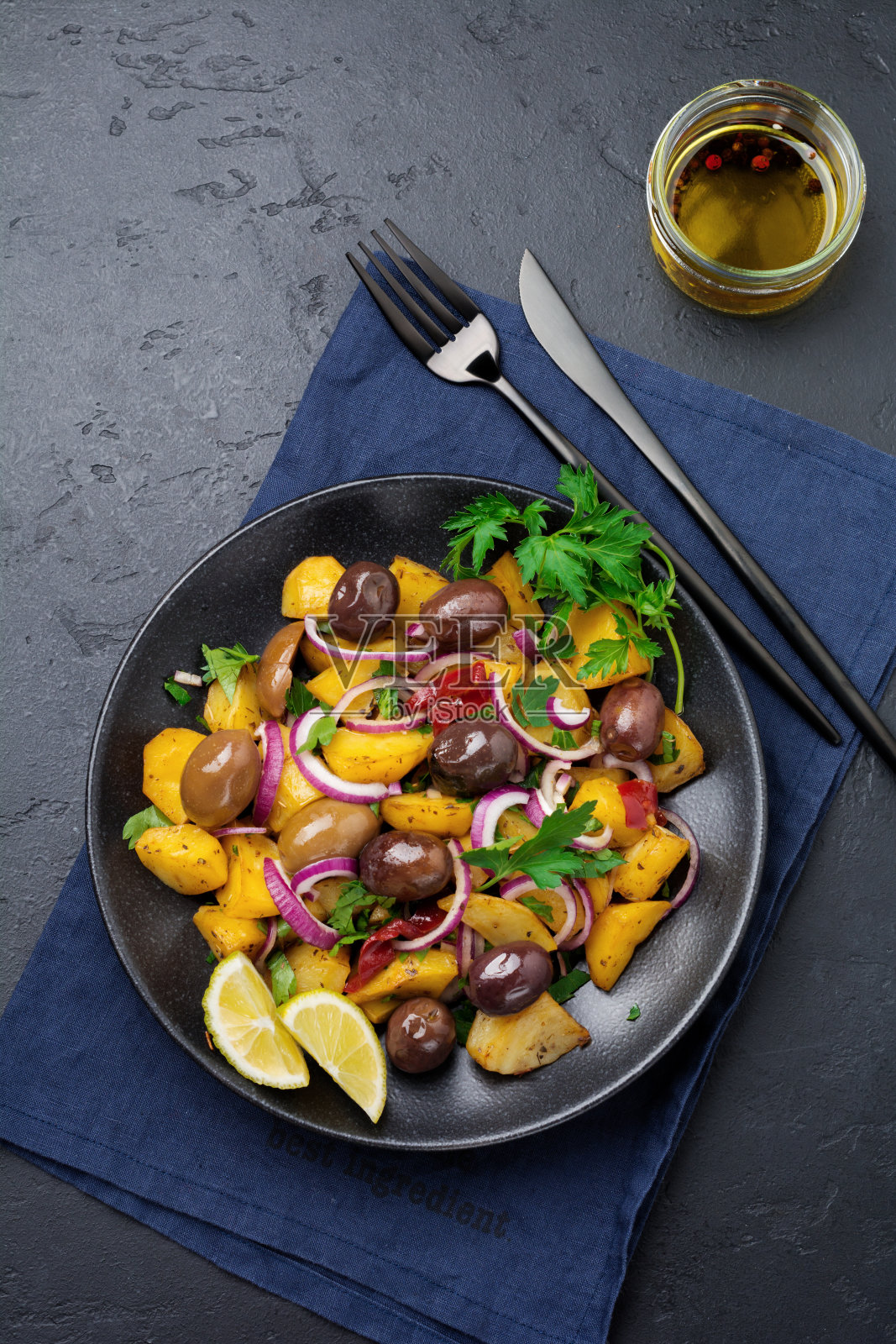 在黑色陶瓷盘子上放上热土豆沙拉，配上橄榄、胡椒、欧芹和红洋葱，背景是黑色的混凝土。有选择性的重点。前视图。副本的空间。照片摄影图片