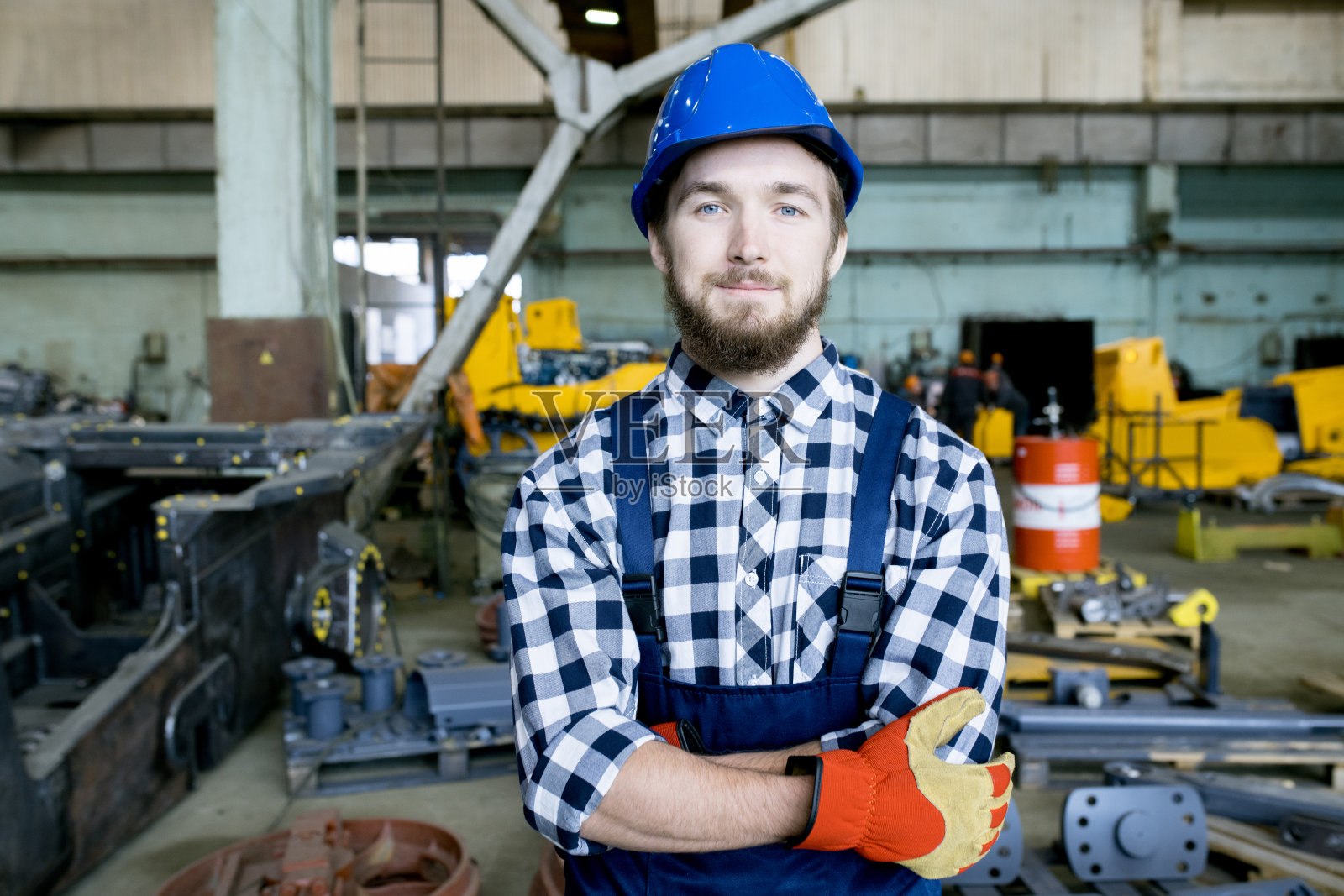 内容英俊的年轻手工工人拖拉机厂戴着蓝色安全帽交叉在胸前，看着相机在工业车间照片摄影图片