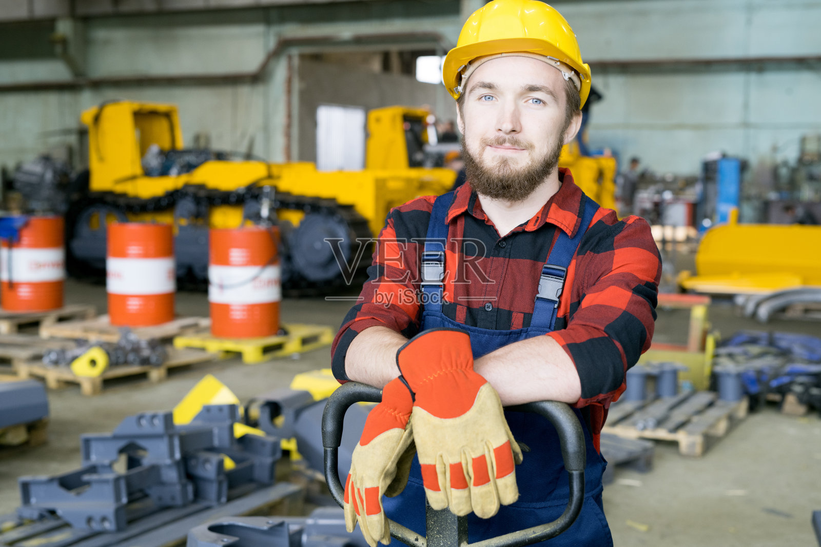 内容英俊的年轻留胡子的搬运工，戴着黄色安全帽，戴着手套，双手倚靠在托盘卡车把手，看着在工业仓库的摄像头照片摄影图片