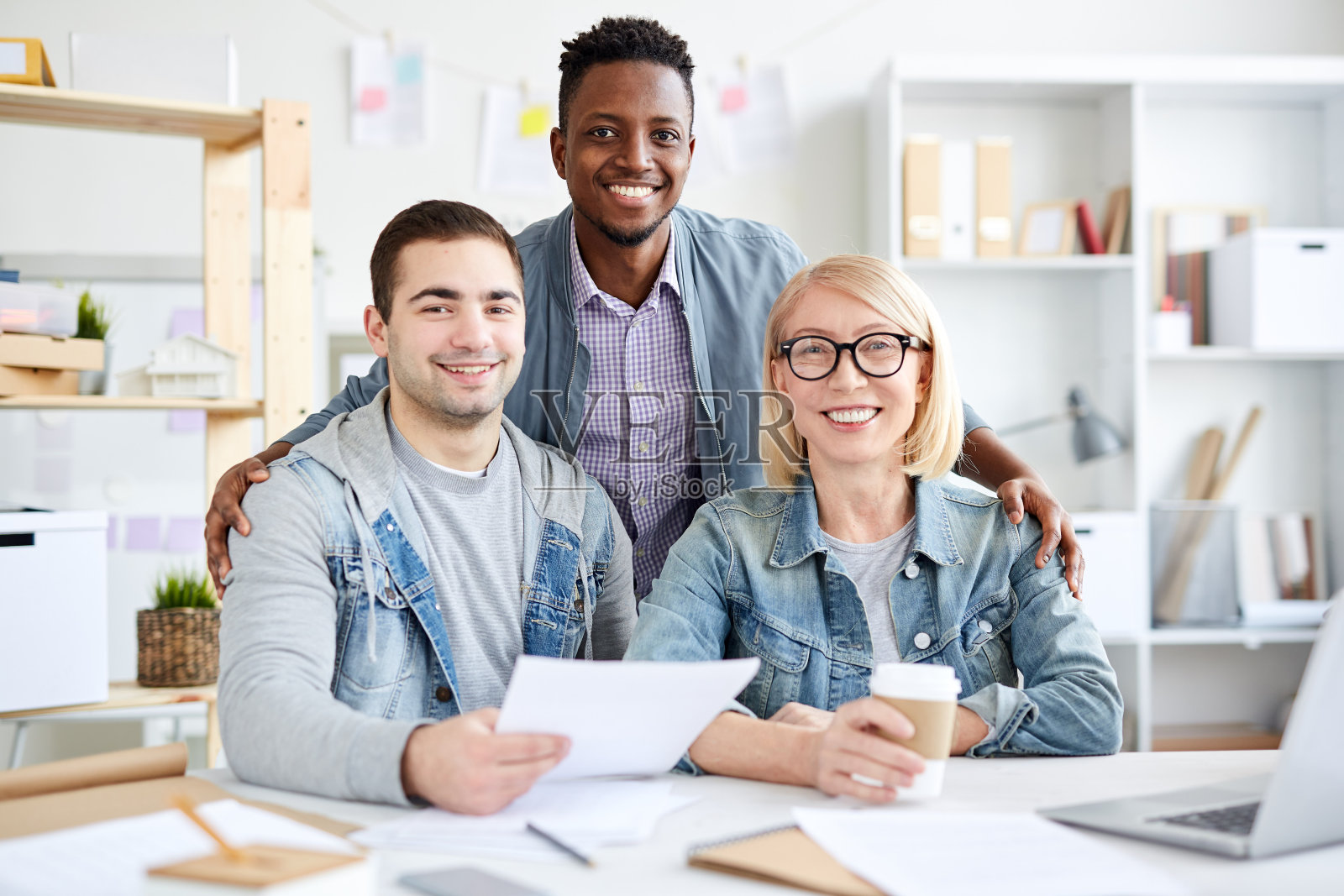 开朗友好的多民族设计师团队对着镜头微笑，英俊的黑人在办公室拥抱同事照片摄影图片