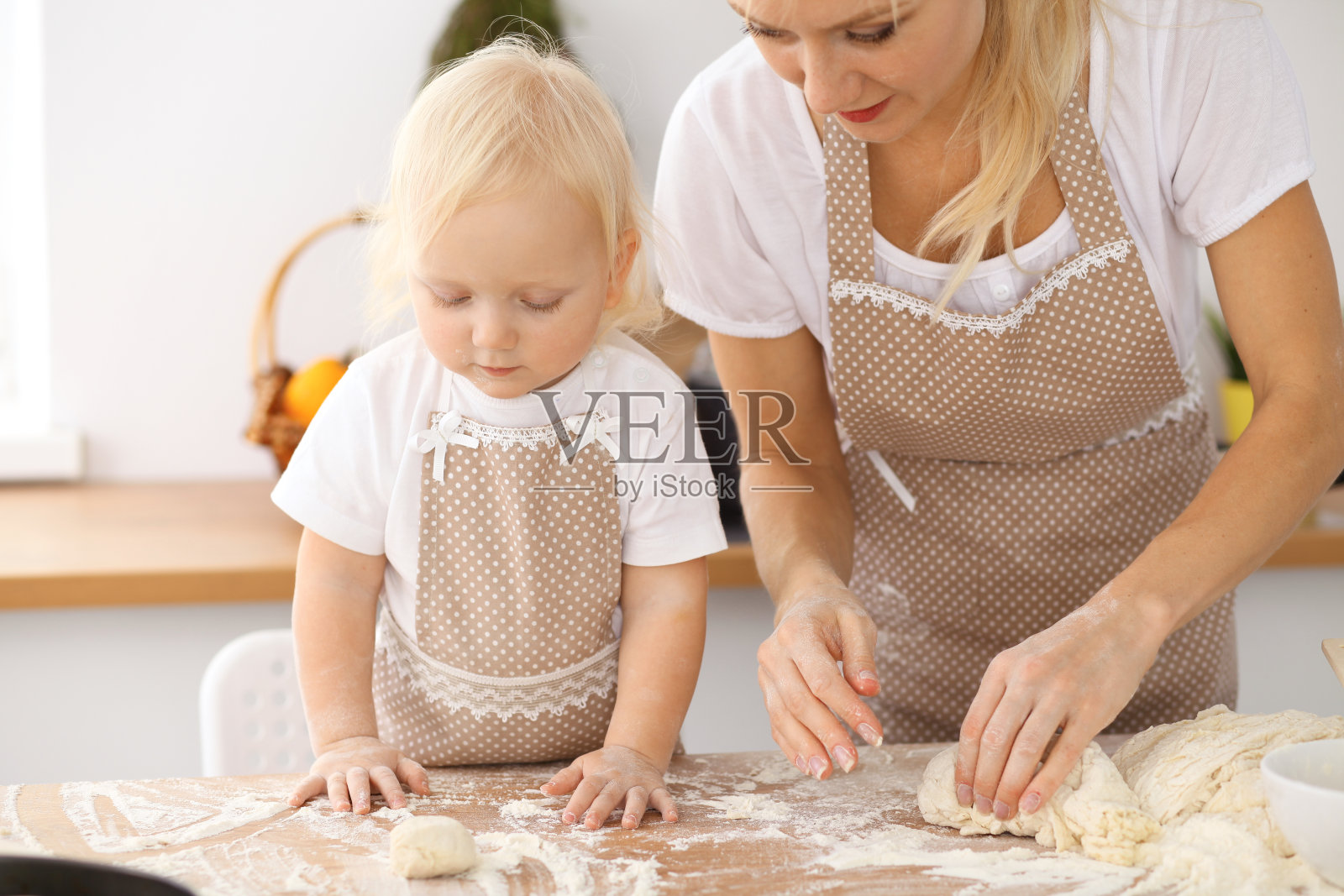 小女孩和她金发碧眼的妈妈穿着米色围裙，一边在厨房揉面，一边又玩又笑。自制糕点，面包，比萨饼或烤饼干照片摄影图片