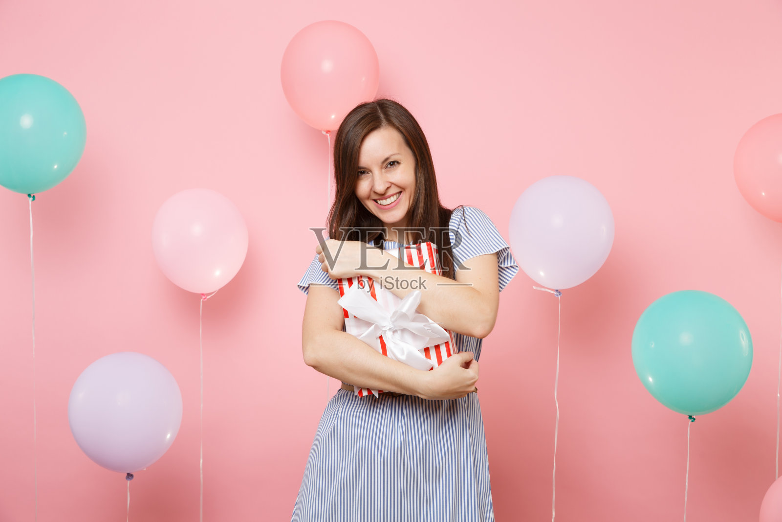 美丽的微笑年轻女子的肖像在蓝色的裙子抱着和拥抱红色盒子的礼物在粉红色的背景与彩色的气球。生日节日聚会，让人真挚情感。照片摄影图片