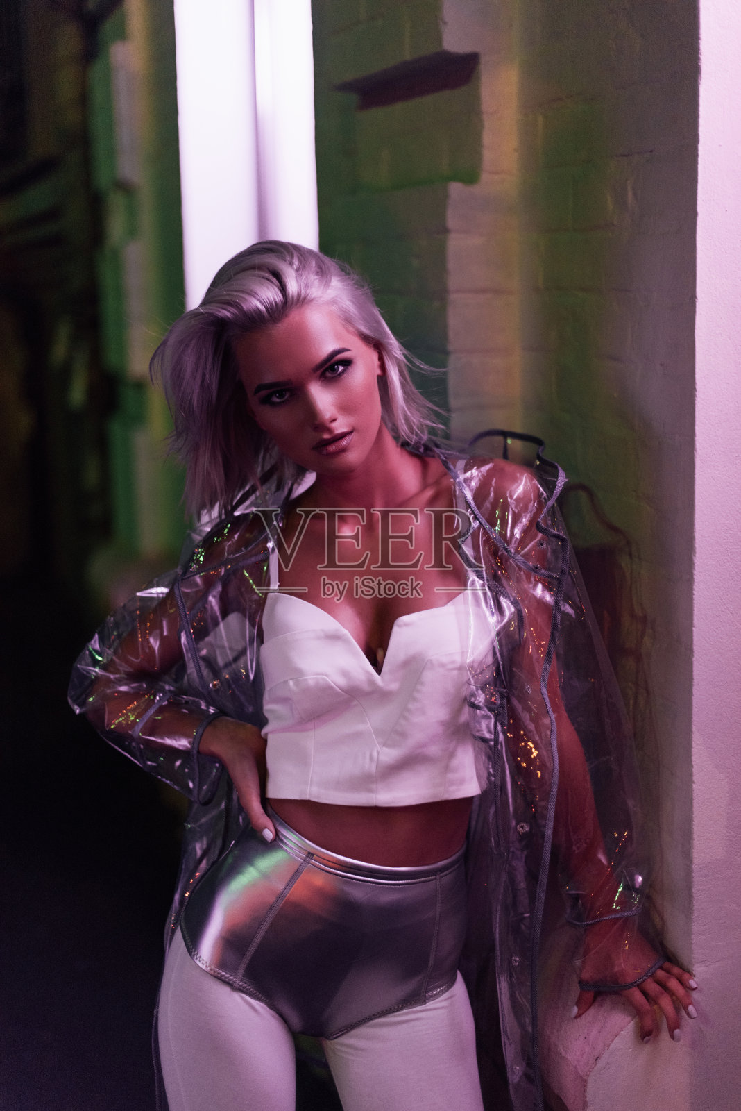 迷人的年轻女子穿着透明的雨衣站在夜晚的街道上，在粉红的灯光下照片摄影图片