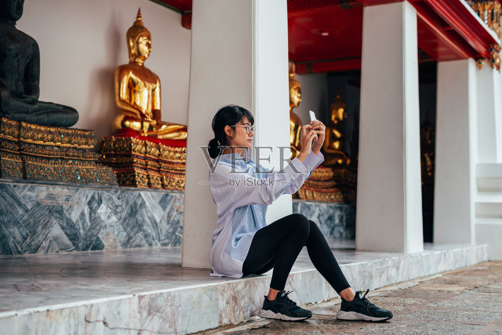 亚洲女性游客坐着用智能手机自拍，以古老的佛像为背景——泰国曼谷的卧佛寺——亚洲。照片摄影图片