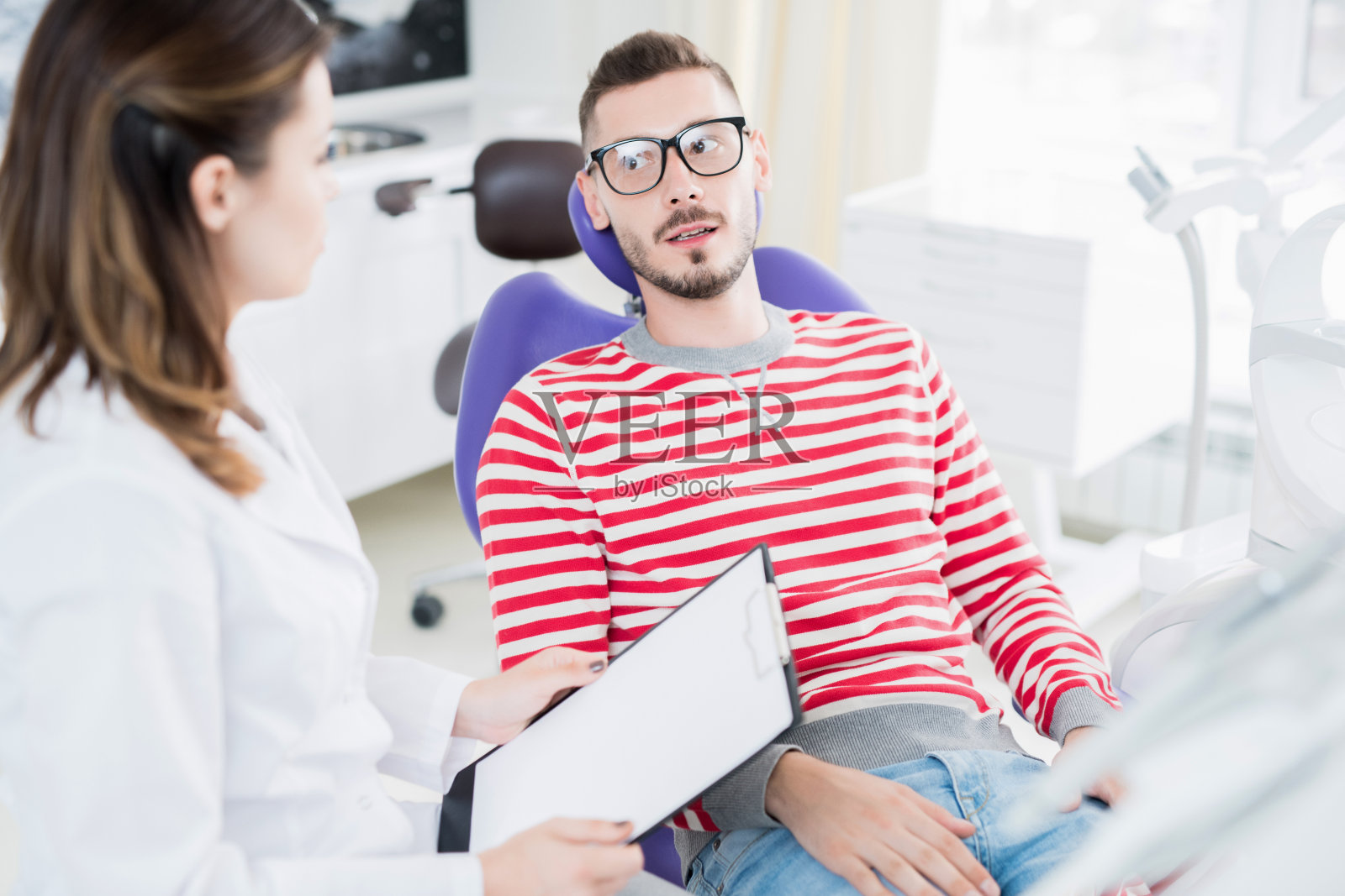 冷静英俊的年轻男子在脱衣坐在牙科椅子上，看着牙医，而告诉牙齿问题在诊所访问照片摄影图片