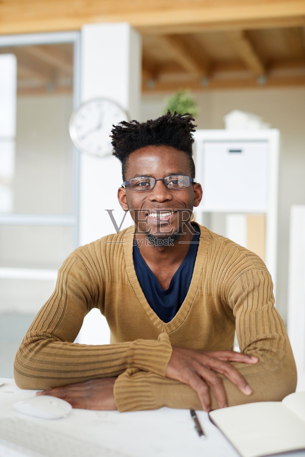 积极兴奋的年轻非洲人与辫穿着时尚的毛衣坐在桌子上，看着办公室的相机照片摄影图片