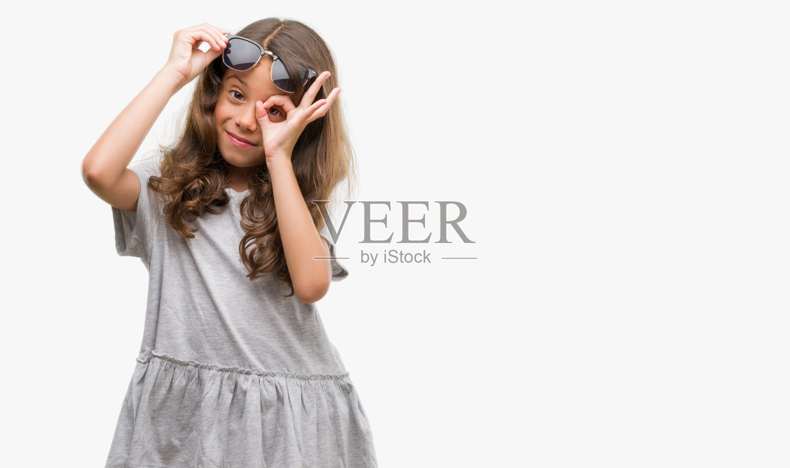 黑发的西班牙裔女孩戴着太阳镜做着ok的手势，手微笑，眼睛透过手指看着幸福的脸。照片摄影图片