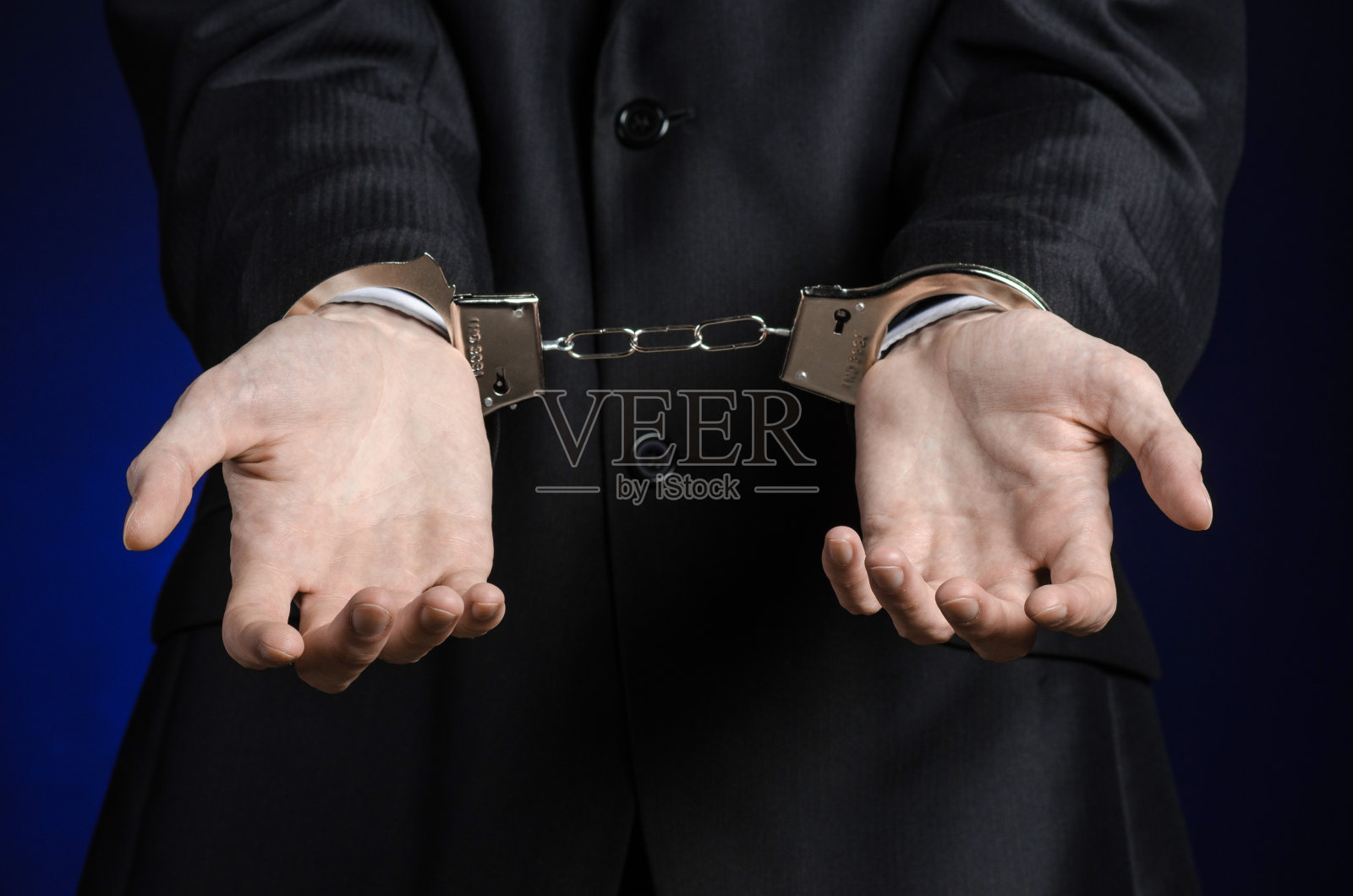 腐败与贿赂主题:商人身穿黑色西装，双手戴着手铐，以深蓝色为背景，在演播室孤立照片摄影图片