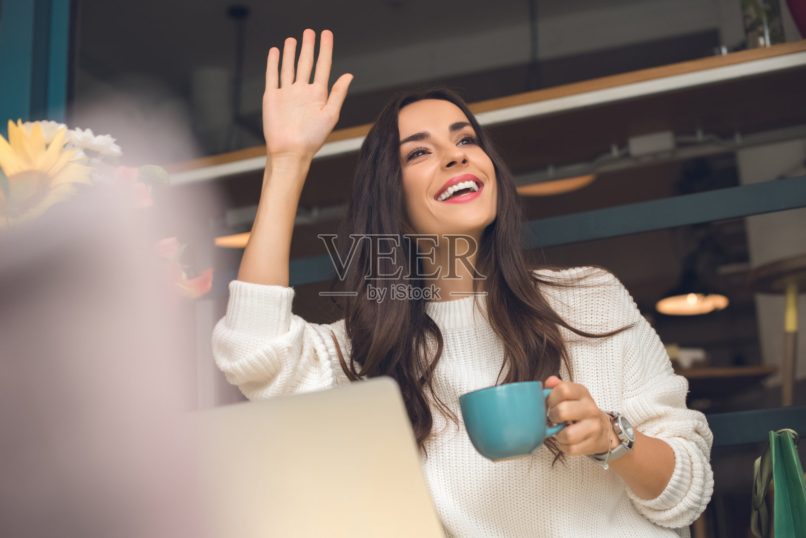 快乐的女性自由职业者与咖啡杯手挥手在桌子上与笔记本电脑在咖啡馆照片摄影图片