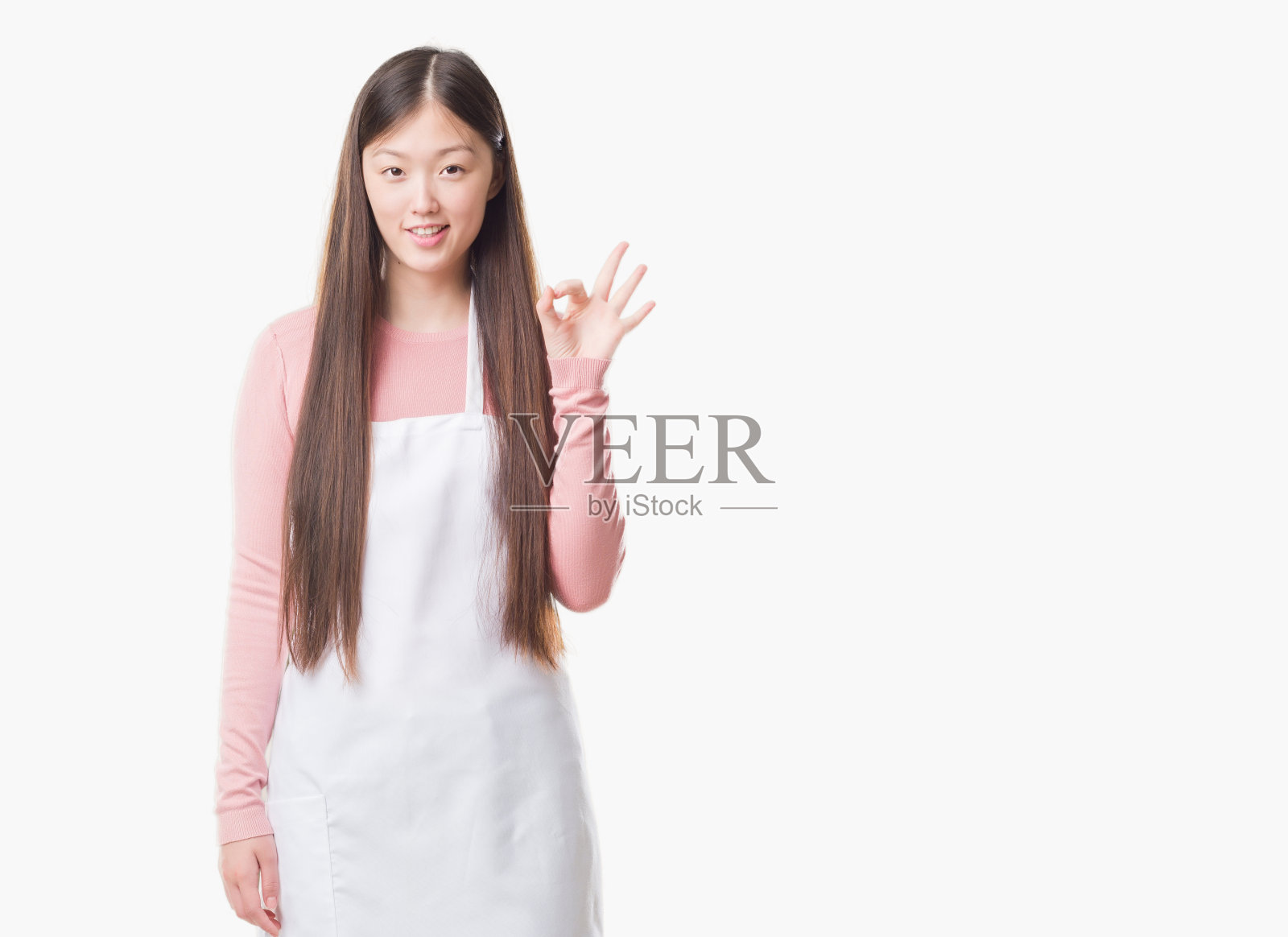 年轻的中国女店主在孤立的背景下，穿着白色围裙，用手指做ok的手势，很好的象征照片摄影图片