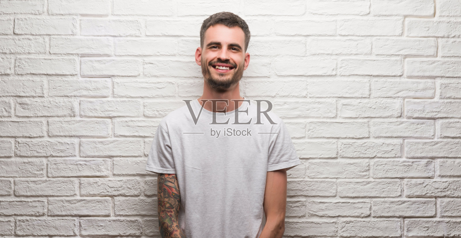 一个年轻的成年人站在白色的砖墙上，脸上带着快乐的表情站着，脸上带着自信的微笑露出牙齿照片摄影图片