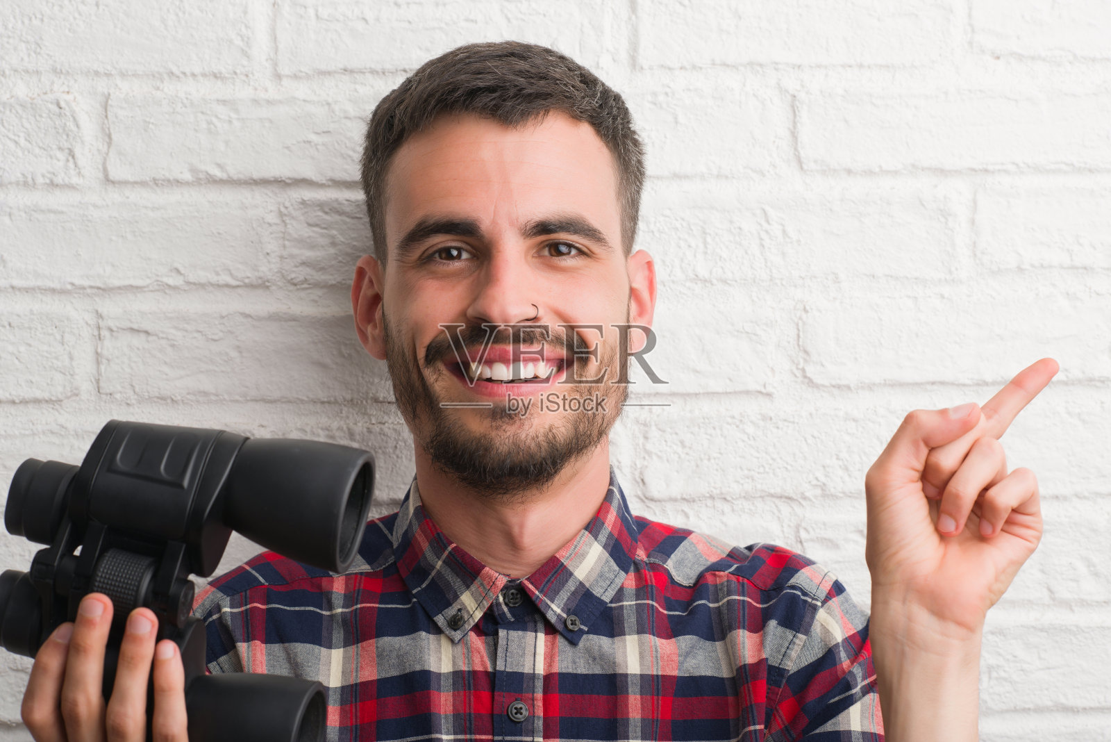年轻的成年男子越过砖墙透过双筒望远镜很高兴地用手和手指指着一边照片摄影图片