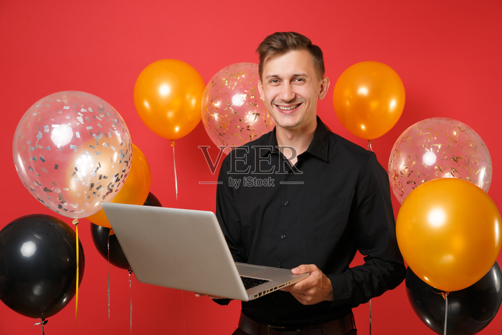 英俊的年轻人在黑色经典衬衫拿着笔记本电脑电脑在明亮的红色背景的气球。情人节，国际妇女节新年生日模型节日派对概念。照片摄影图片