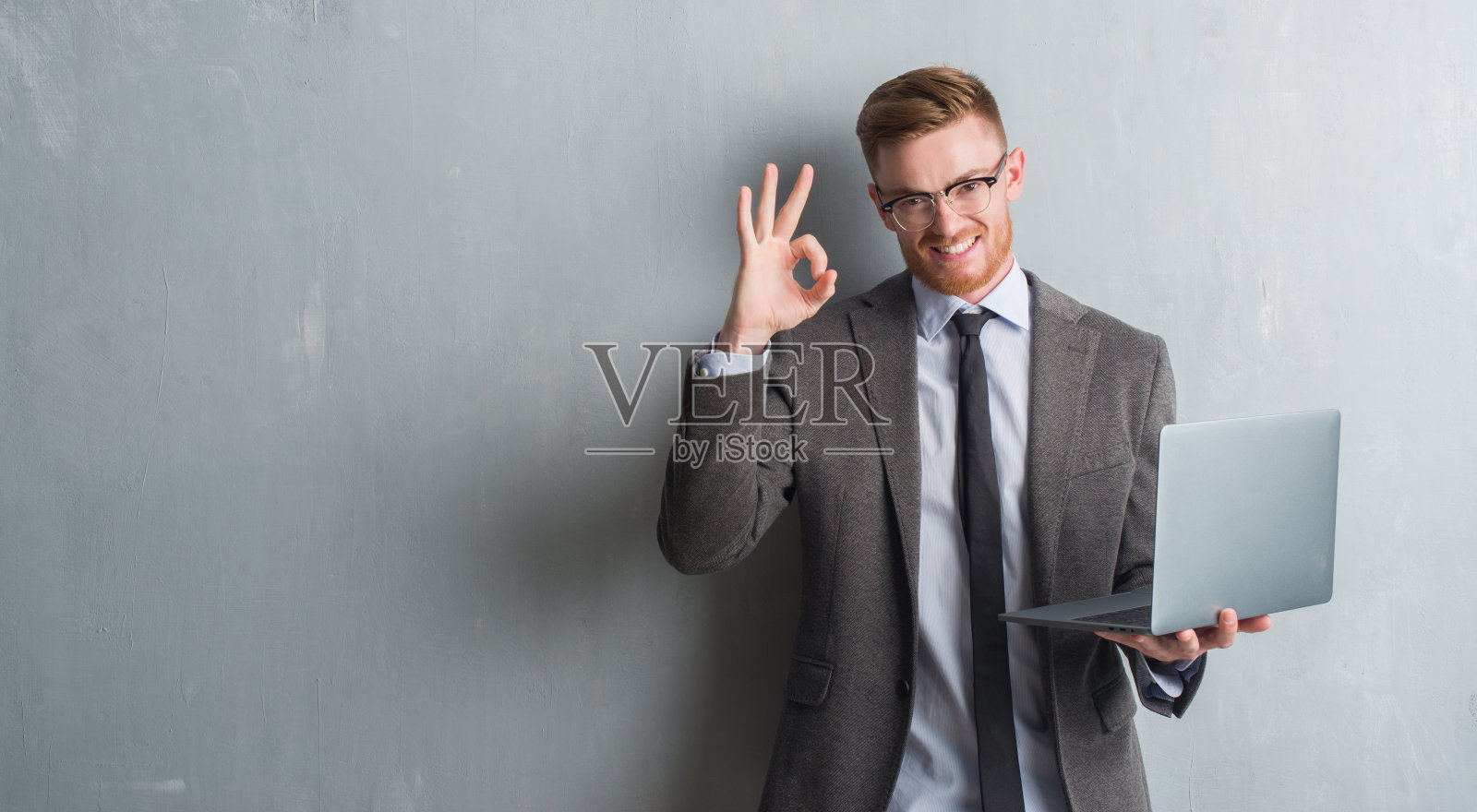 年轻的红头发优雅的商人在灰色的垃圾墙用手提电脑做ok手势的手指，优秀的象征照片摄影图片