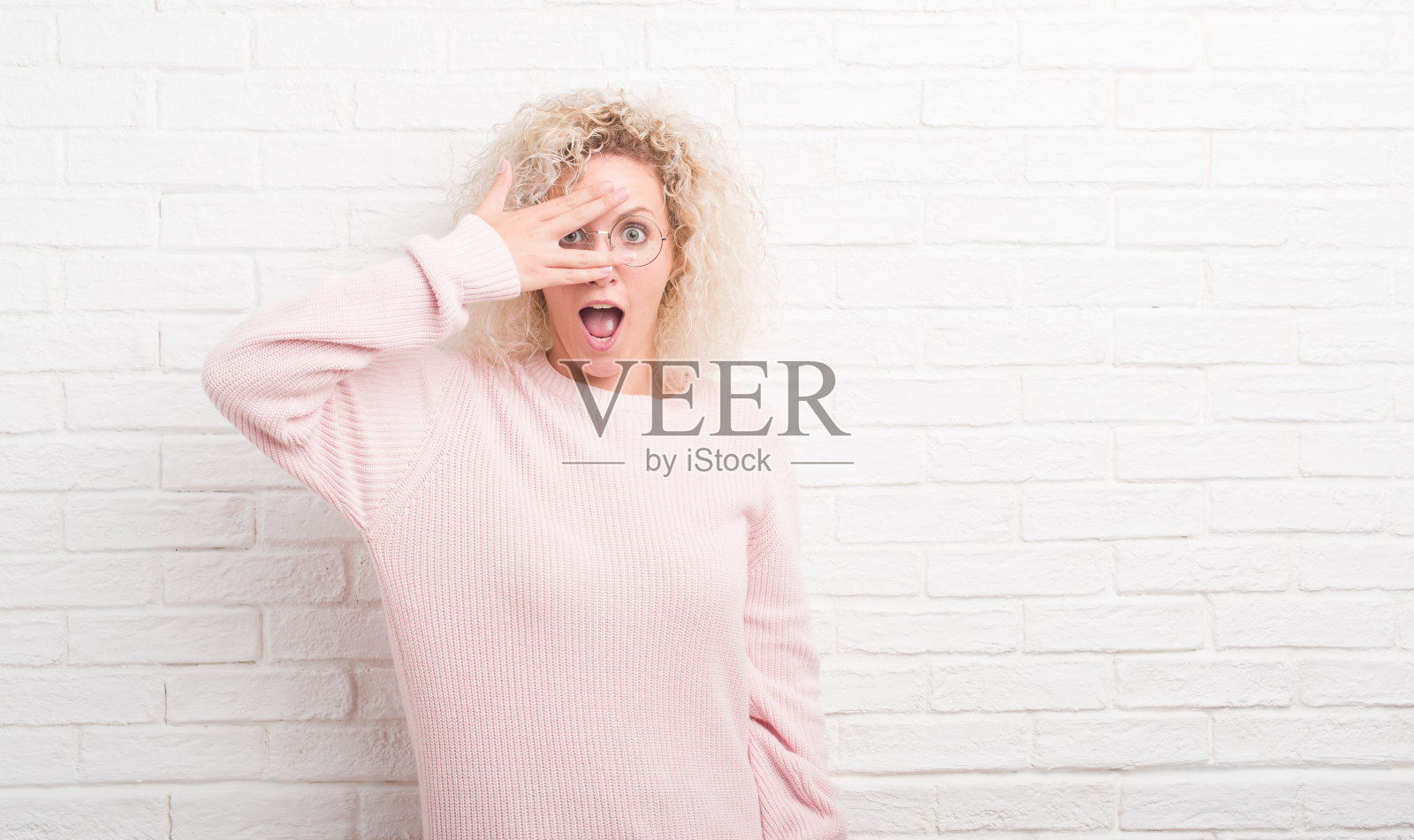 年轻的金发女郎，一头卷发，越过白色的砖墙，用手捂着脸和眼睛，在震惊中窥视，透过手指，带着尴尬的表情。照片摄影图片