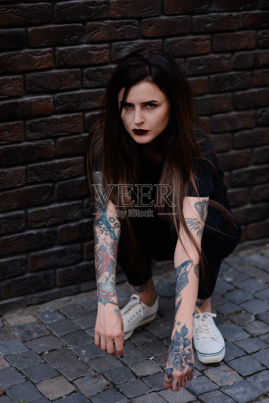 美丽苗条的黑发女孩，黑嘴唇，纹身和穿黑衣服坐在砖墙上照片摄影图片