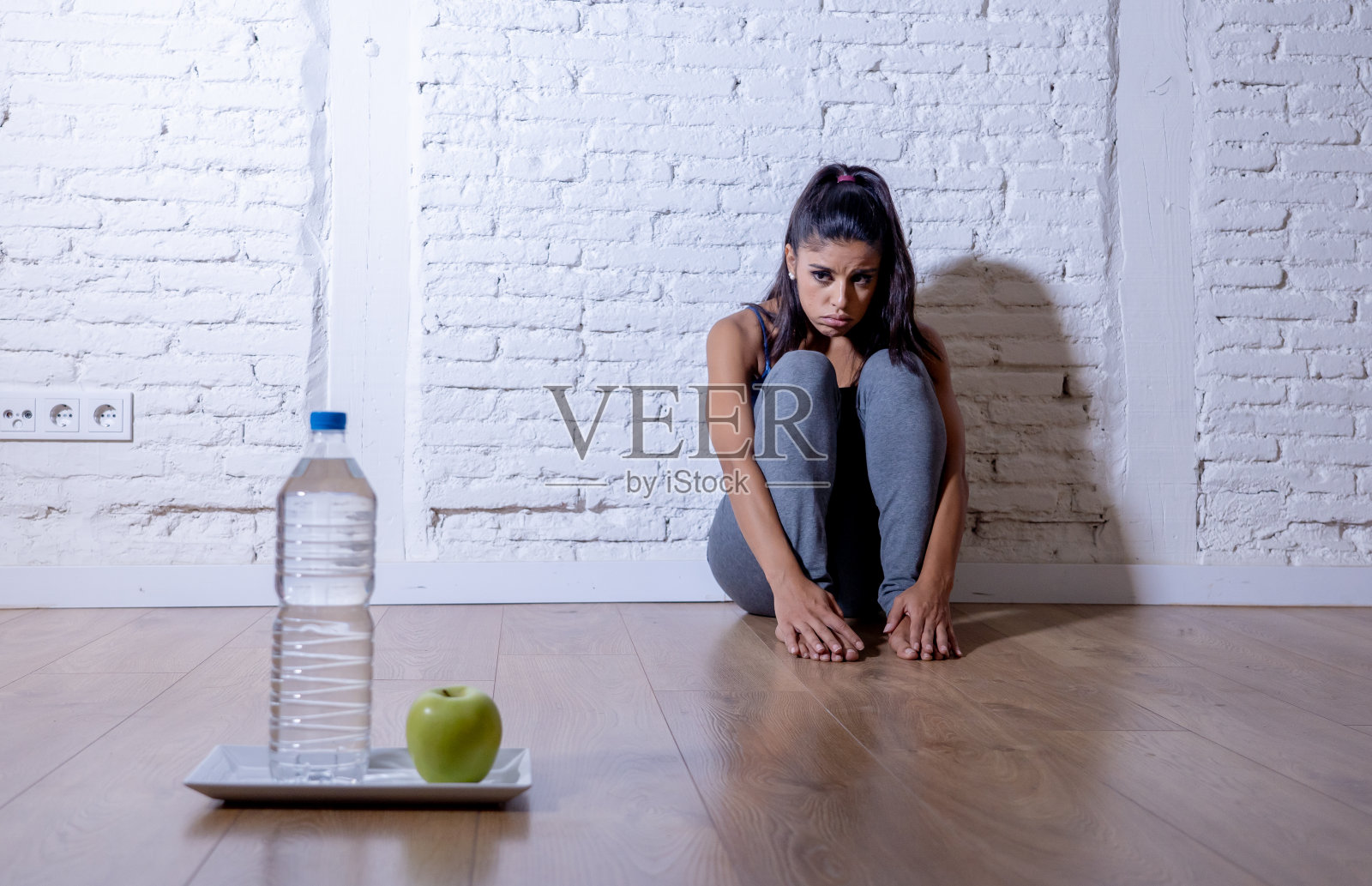 绝望的节食年轻女子坐在地上看着悲伤的一个苹果，水果和水作为疯狂的节食意志力和营养紊乱的象征。照片摄影图片