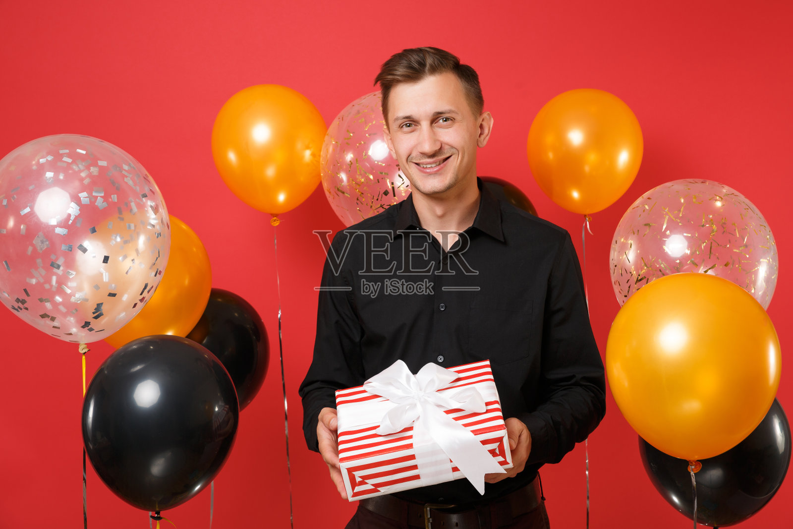 微笑的年轻人在黑色经典衬衫拿着红色盒子的礼物，在明亮的红色背景气球。情人节，妇女节，新年快乐，生日模型节日派对概念。照片摄影图片