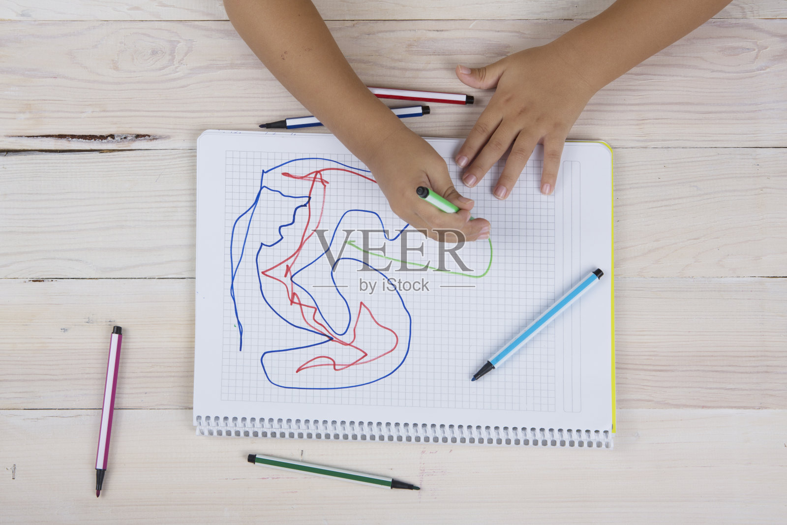 小男孩双手拿着螺旋式记事本和彩色铅笔放在木桌上。照片摄影图片