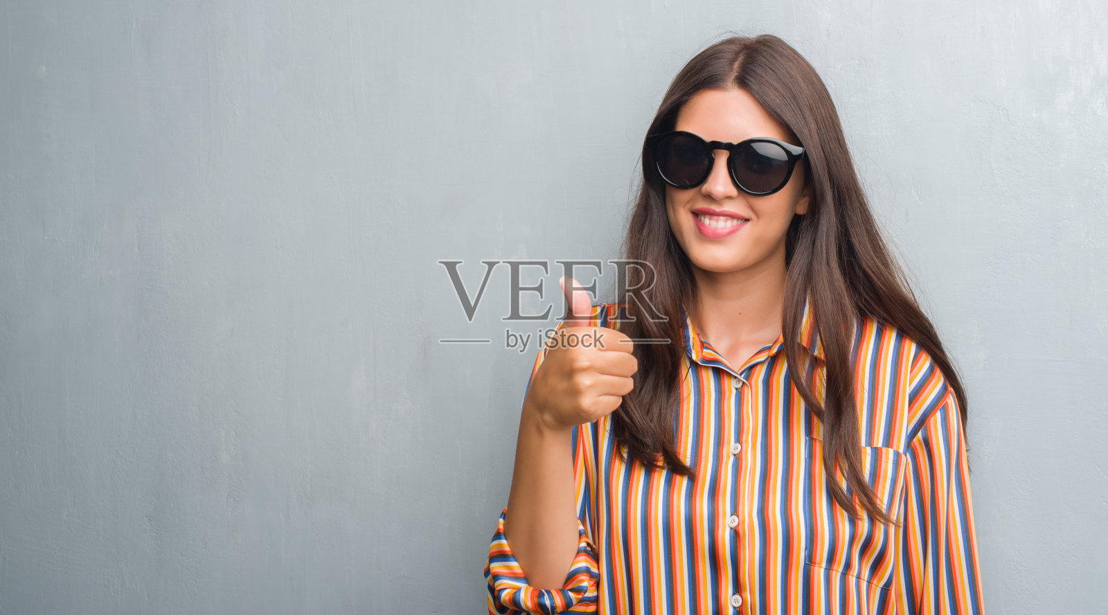年轻的黑发女人越过肮脏的灰色墙壁，戴着大太阳镜，开心地笑着做ok的手势，大拇指向上的手指，优秀的手势照片摄影图片