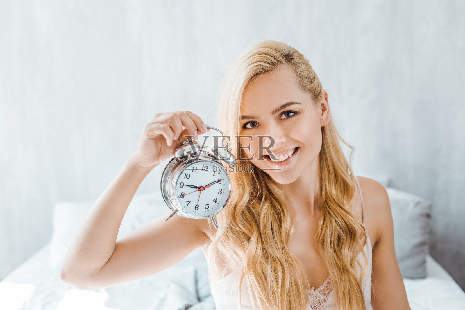 年轻漂亮的白发女性拿着闹钟在卧室里对着相机微笑照片摄影图片