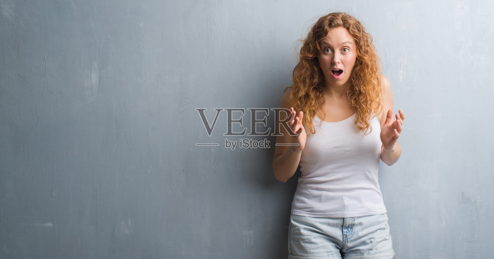 年轻的红发女人越过灰色垃圾墙害怕在震惊与惊讶的脸，害怕和兴奋与恐惧的表情照片摄影图片