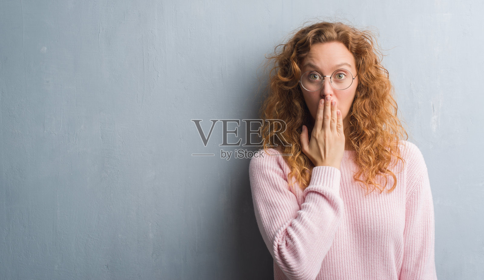 年轻的红发女子越过灰色的垃圾墙戴着眼镜捂着嘴用手为错误而震惊，表情恐惧，在沉默中害怕，秘密的概念照片摄影图片