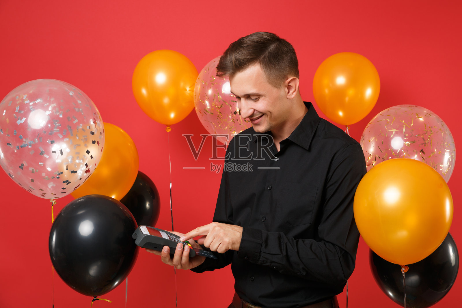 年轻男子手持无线现代银行支付终端进行处理，获取信用卡支付，黑卡上有红色背景的气球。新年生日模型节日派对概念。照片摄影图片