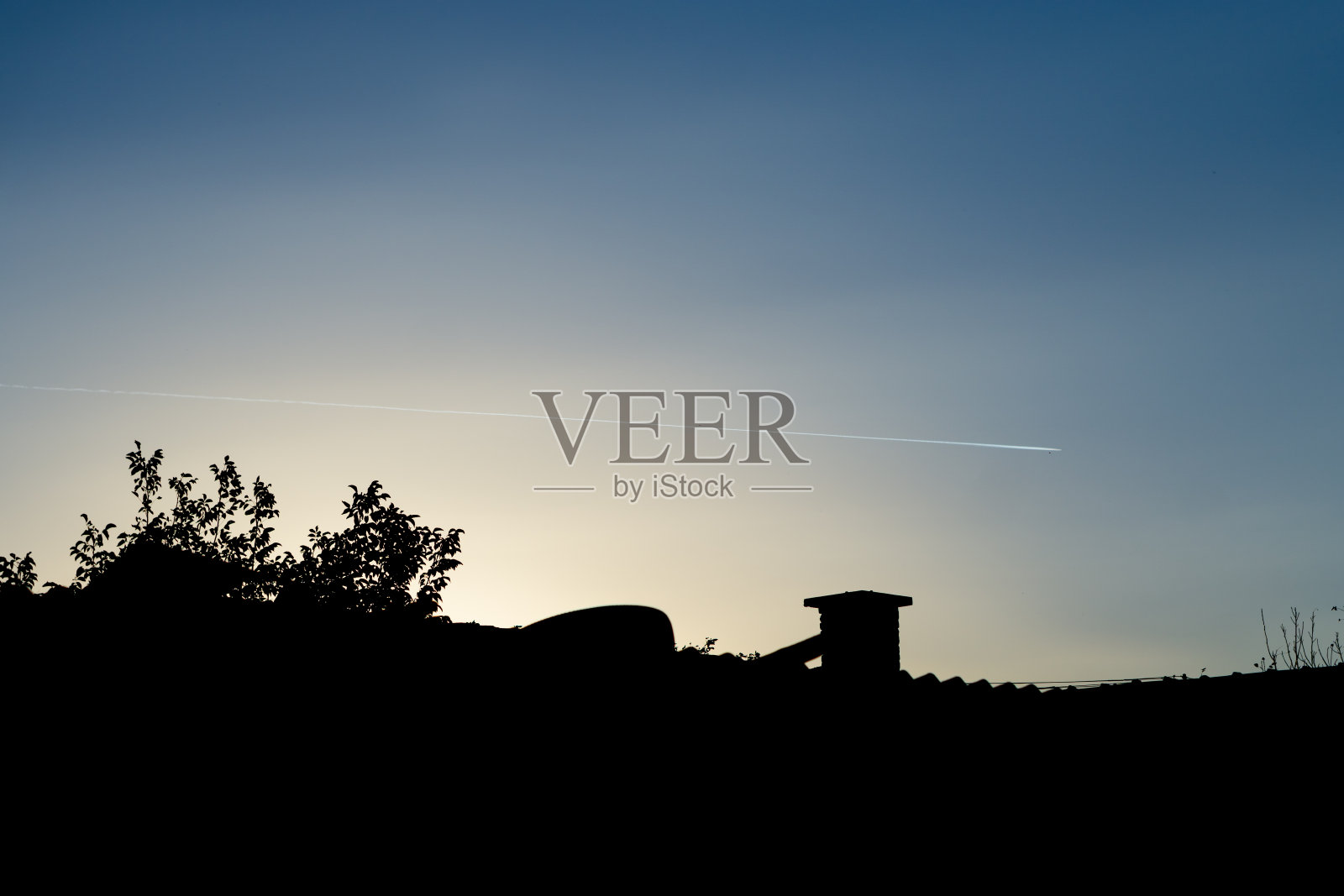 屋顶的剪影与蓝色和黄色的日落天空的背景与飞机的踪迹照片摄影图片