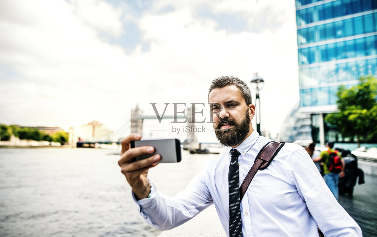 时尚商人拿着智能手机站在伦敦河边自拍。照片摄影图片